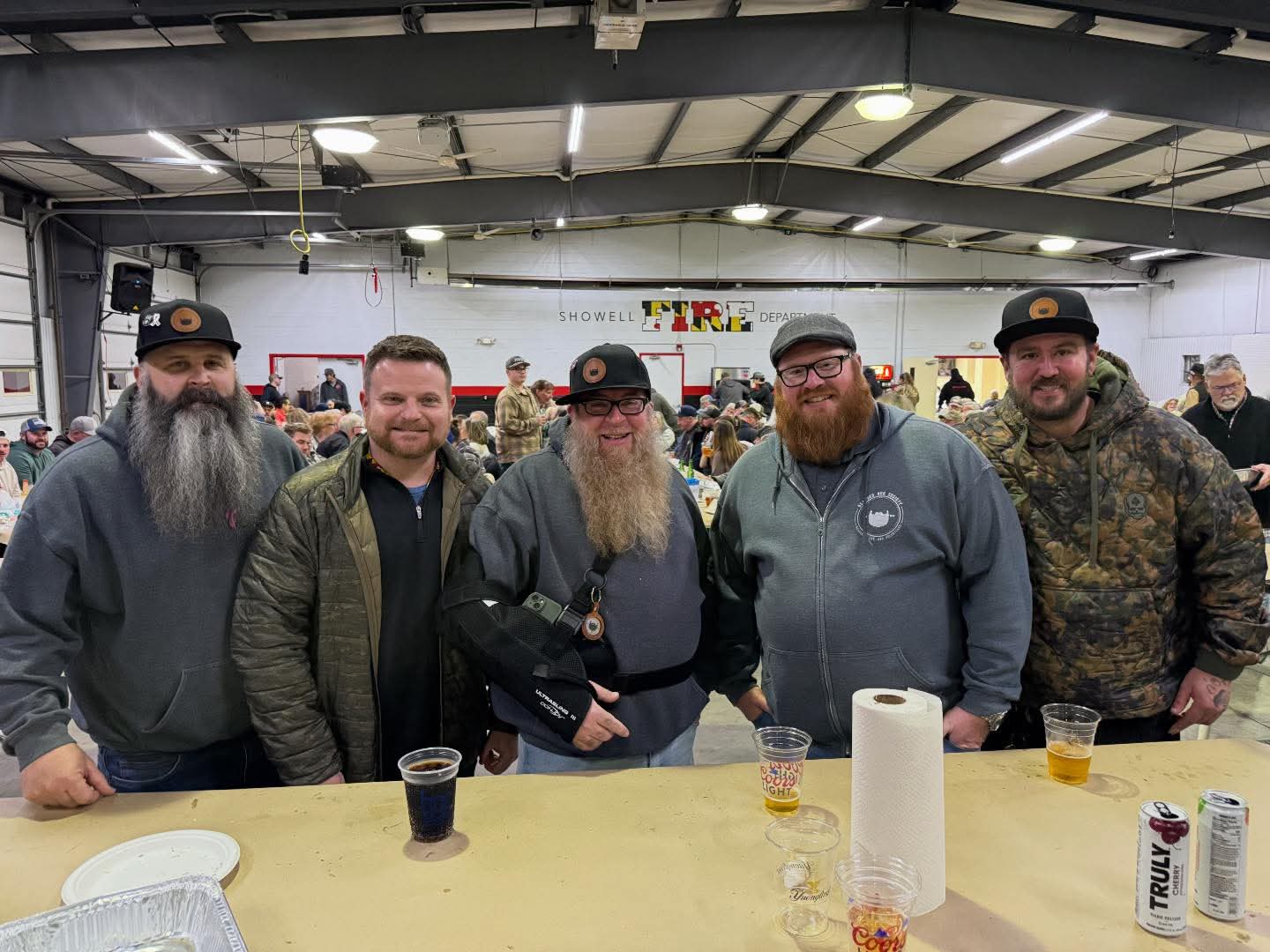 Five men standing behind a table in an indoor setting, some with beards and hats, with drinks and paper towels on the table.