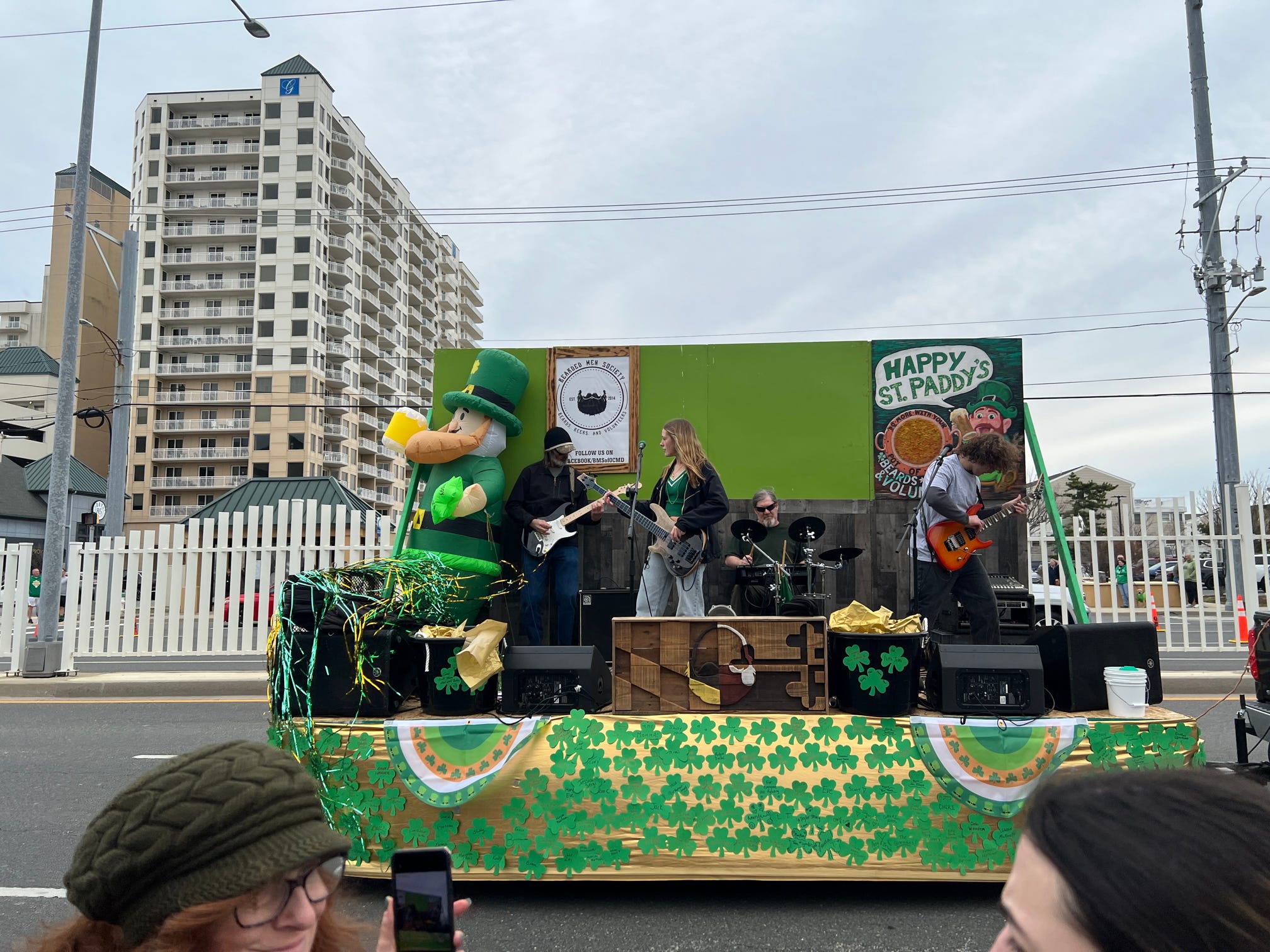 St. Patrick's Day parade float with musicians performing, decorated with shamrocks and leprechaun figure, in an urban area.