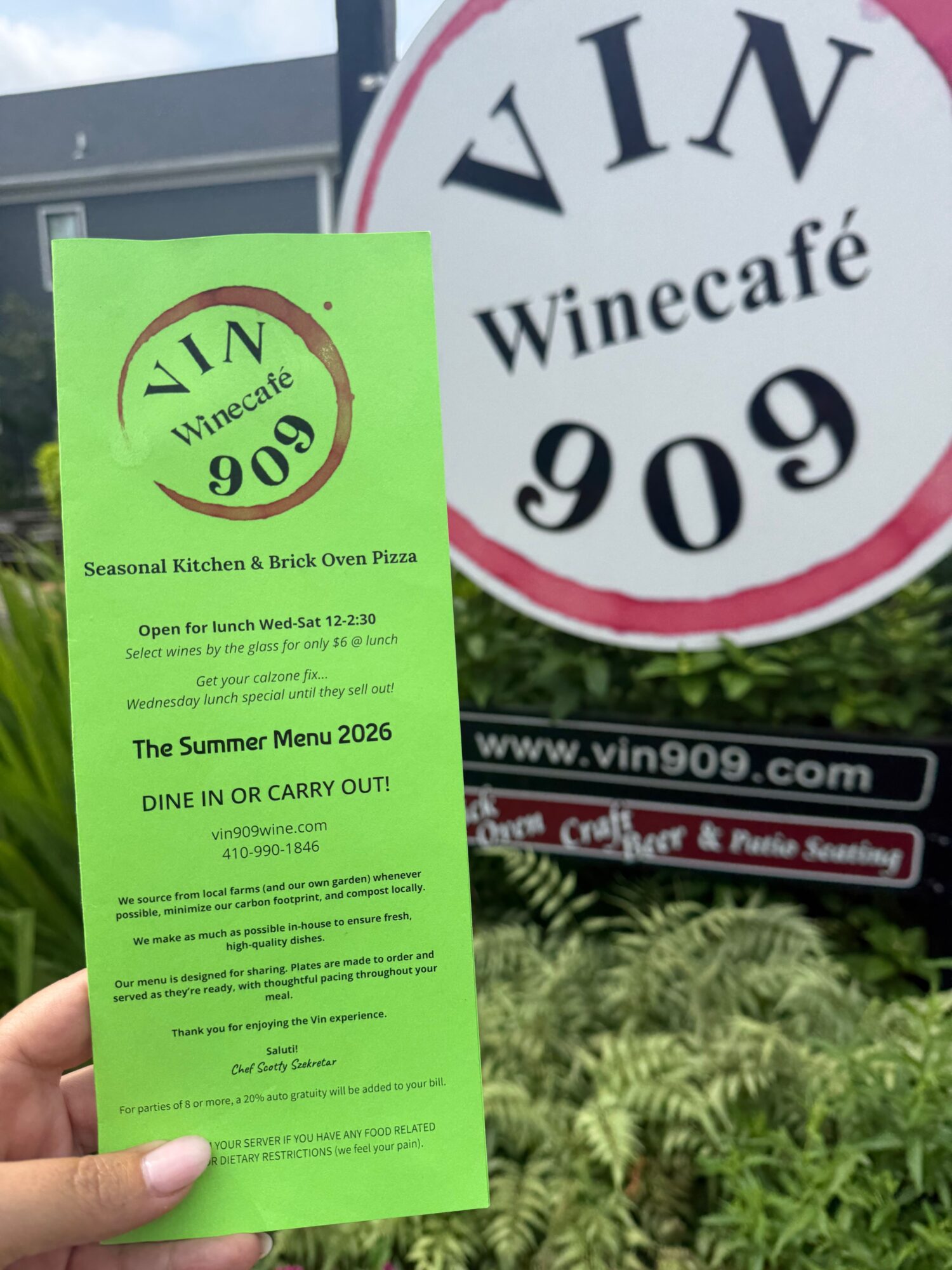 Green ticket with black text and a circular logo, held by a person's hand, with a sign and greenery in the background.