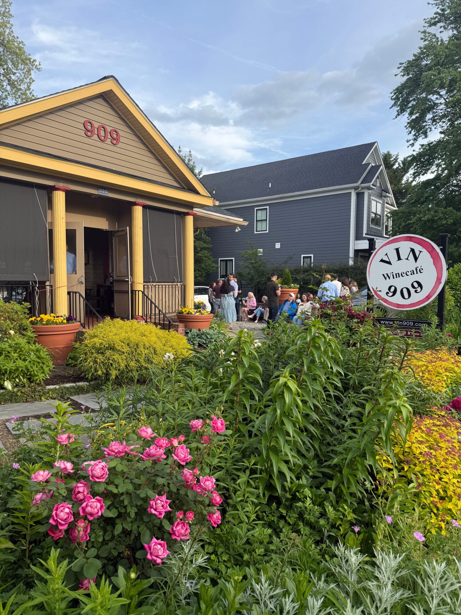 People gather outside a building with a garden of pink flowers and yellow plants, under a cloudy sky.