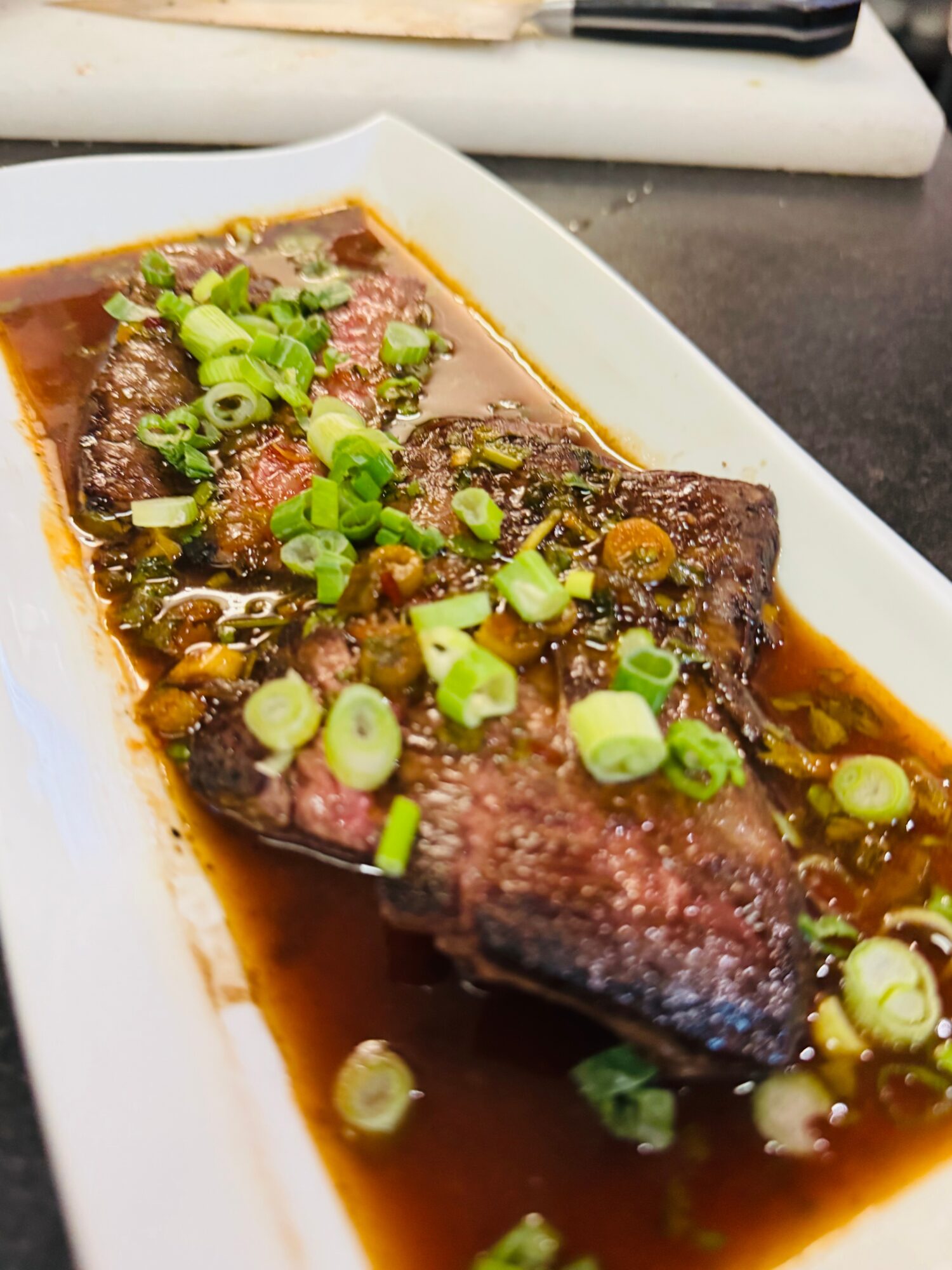 Cooked fish fillet topped with chopped green onions in dark sauce on a white rectangular plate.