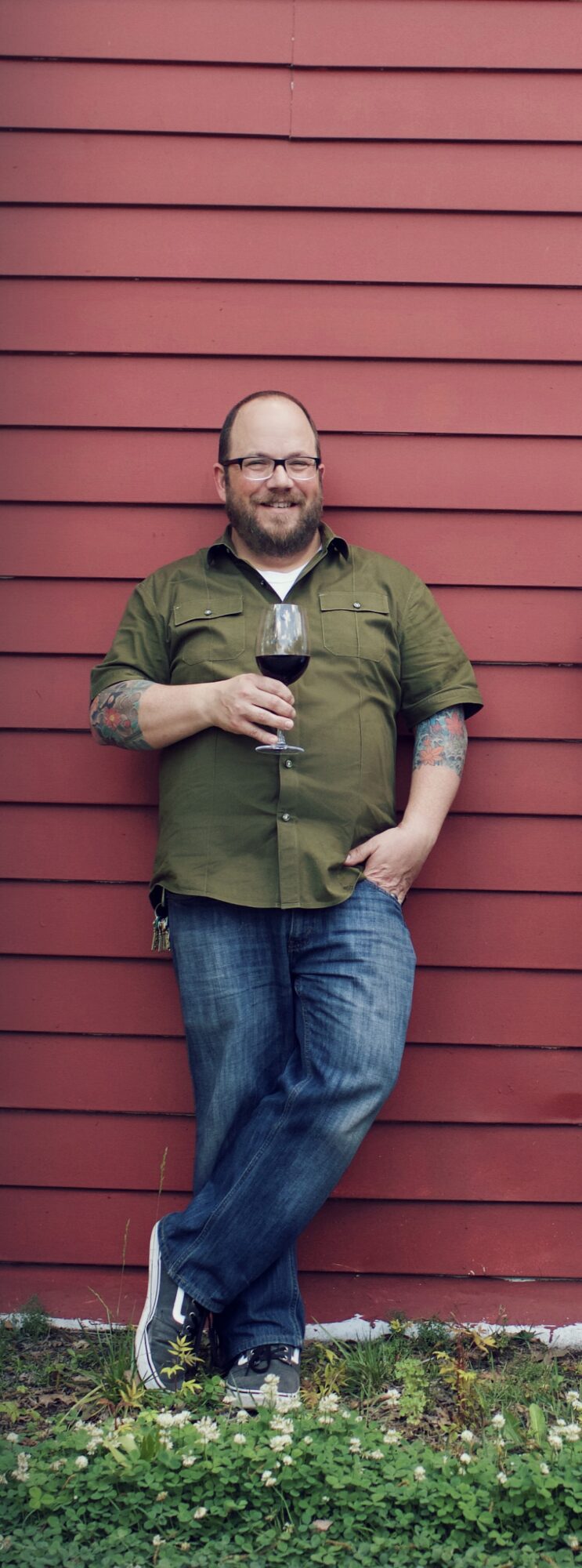 Man with glasses and beard holding a glass of wine, standing against a red wooden wall, smiling.