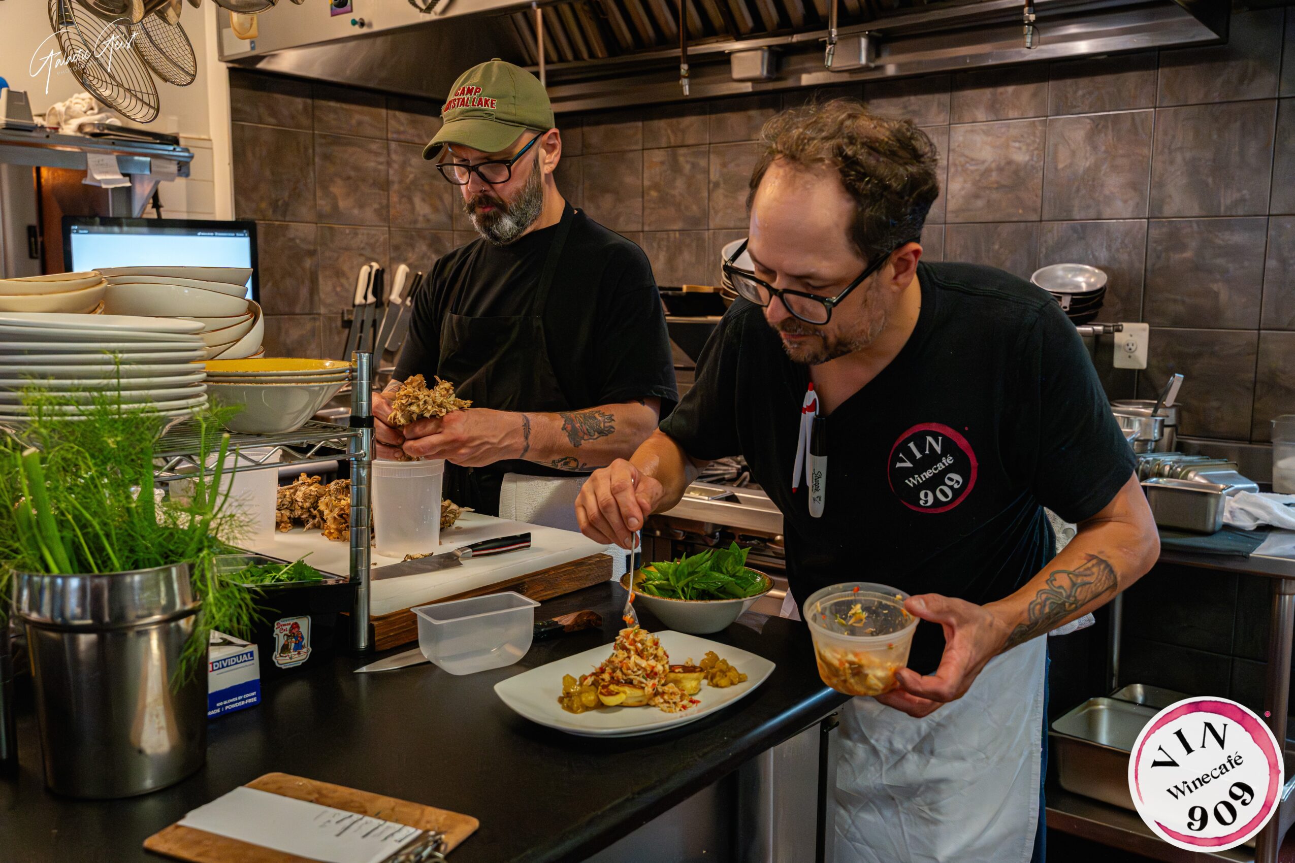Two chefs preparing food in a kitchen, one with a bowl and the other with a plate, both wearing black shirts and glasses.