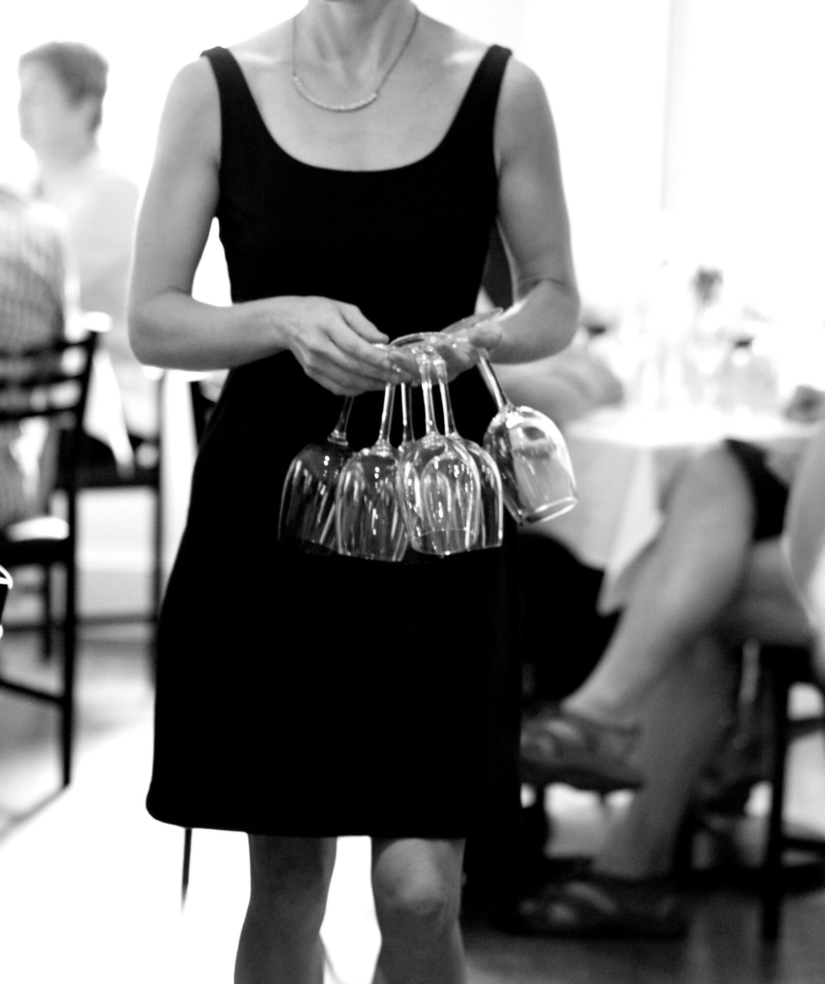 Woman in a black dress holding upside-down wine glasses in a restaurant setting.