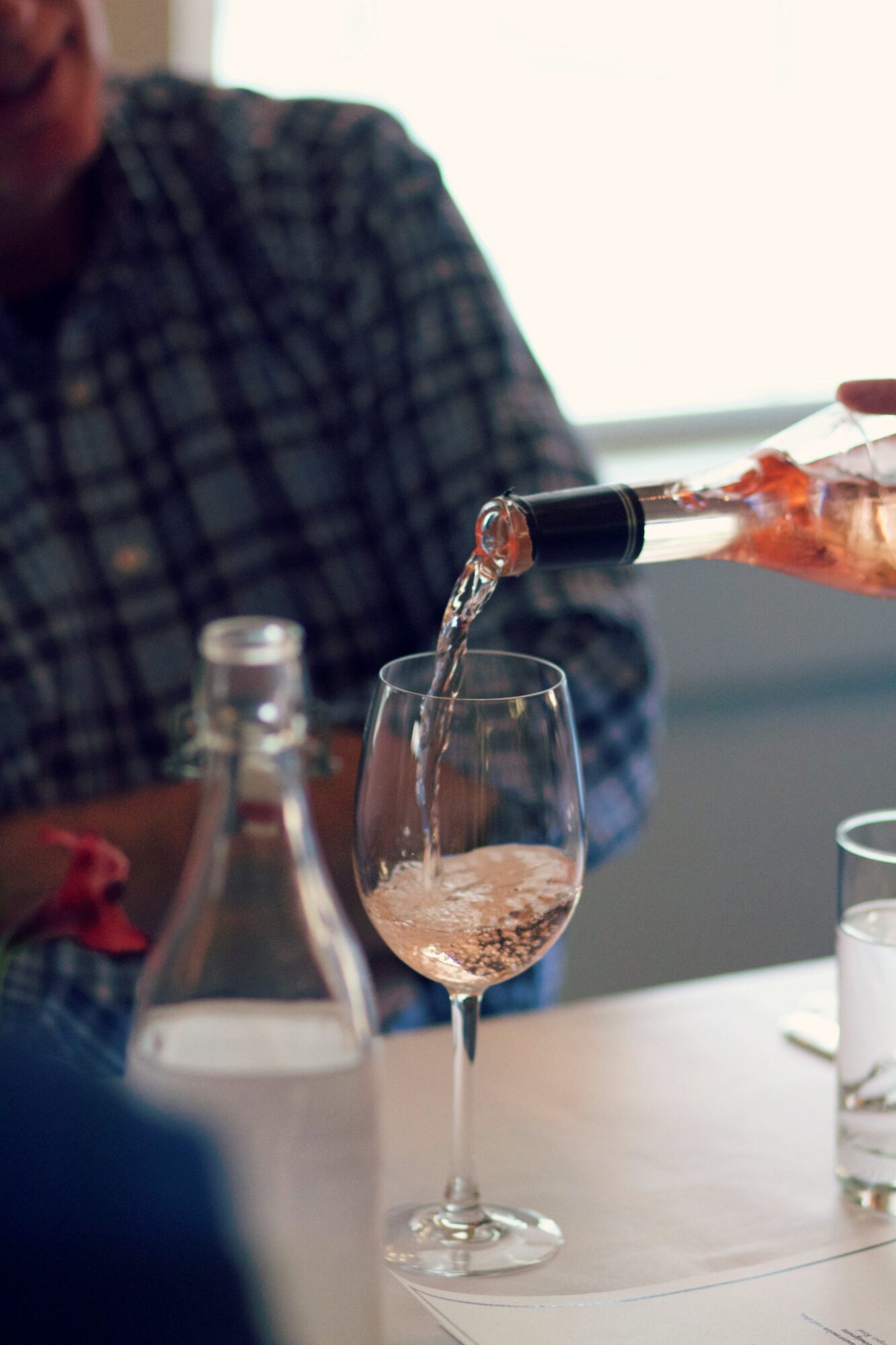 Person pouring rosé wine into a tall glass on a table, with bottles nearby.