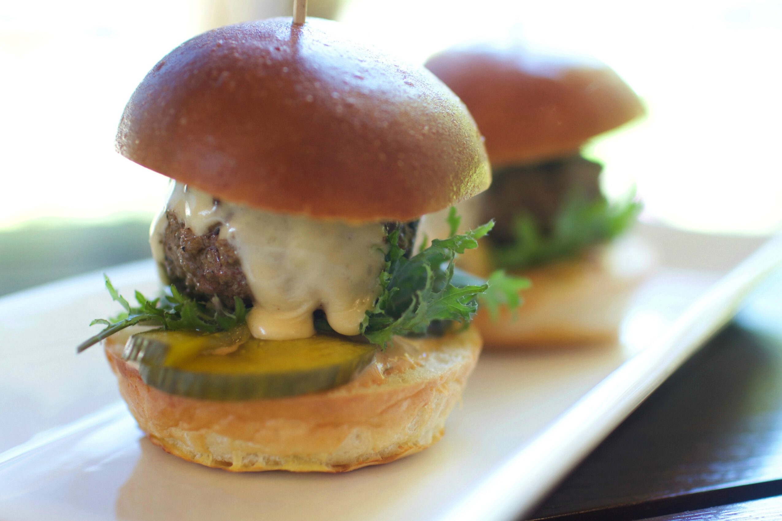 Two mini burgers with pickles, lettuce, and sauce on a white plate, with a blurred background.