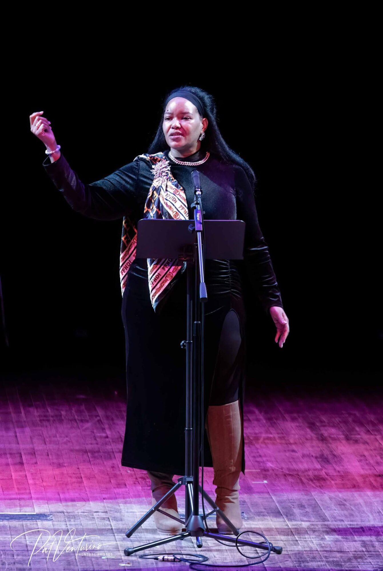 Woman standing on stage, speaking into microphone, gesturing with her right hand, with a dark background and wooden floor.