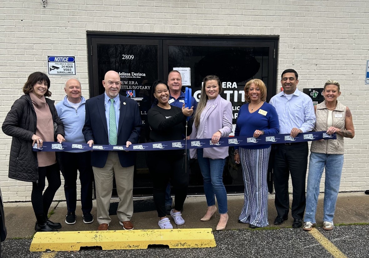 Group of nine people standing outside a building, holding a ribbon for a ribbon-cutting event.