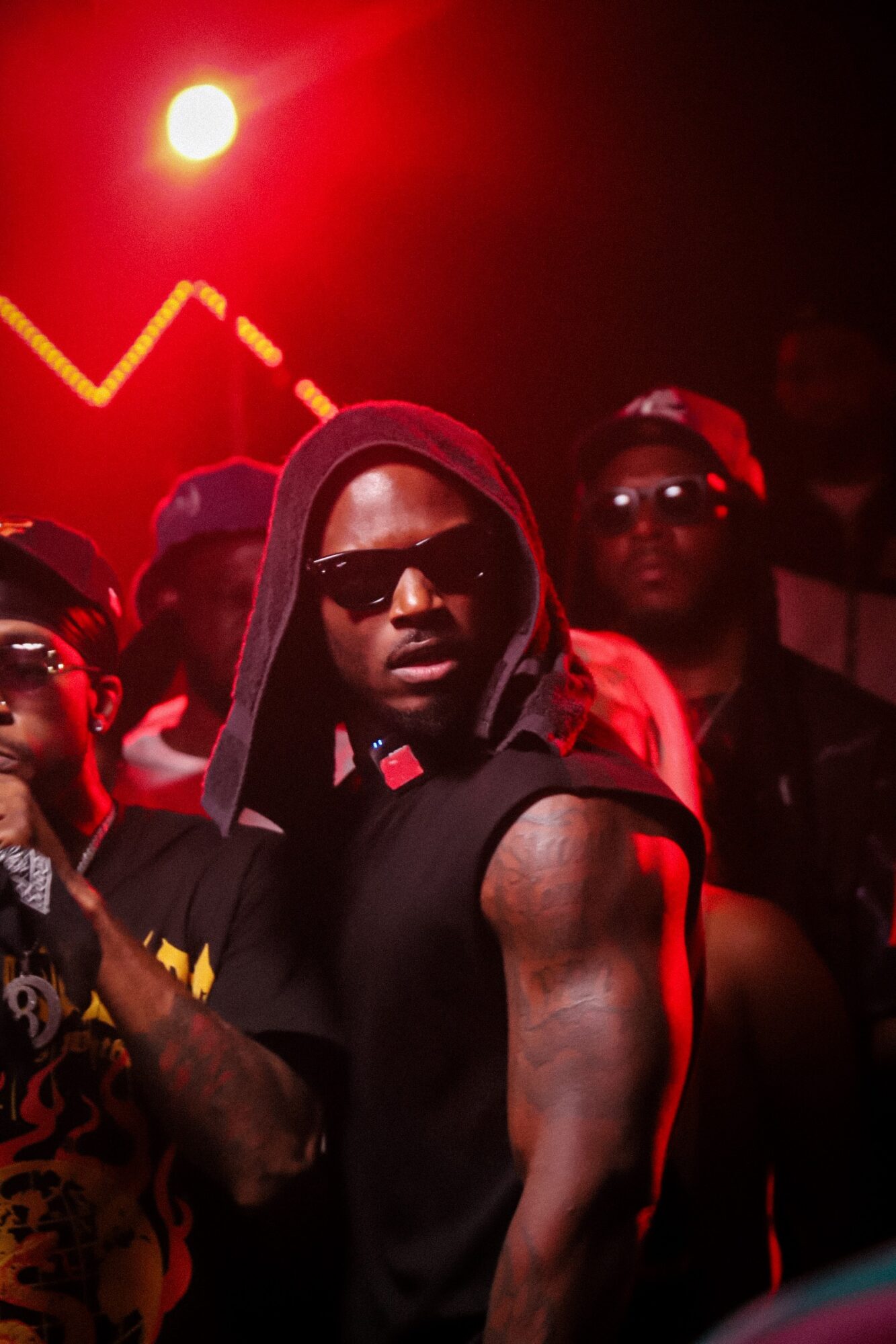 Group of people in dark clothing and sunglasses, with one person in the foreground showing a muscular arm, under red lighting.
