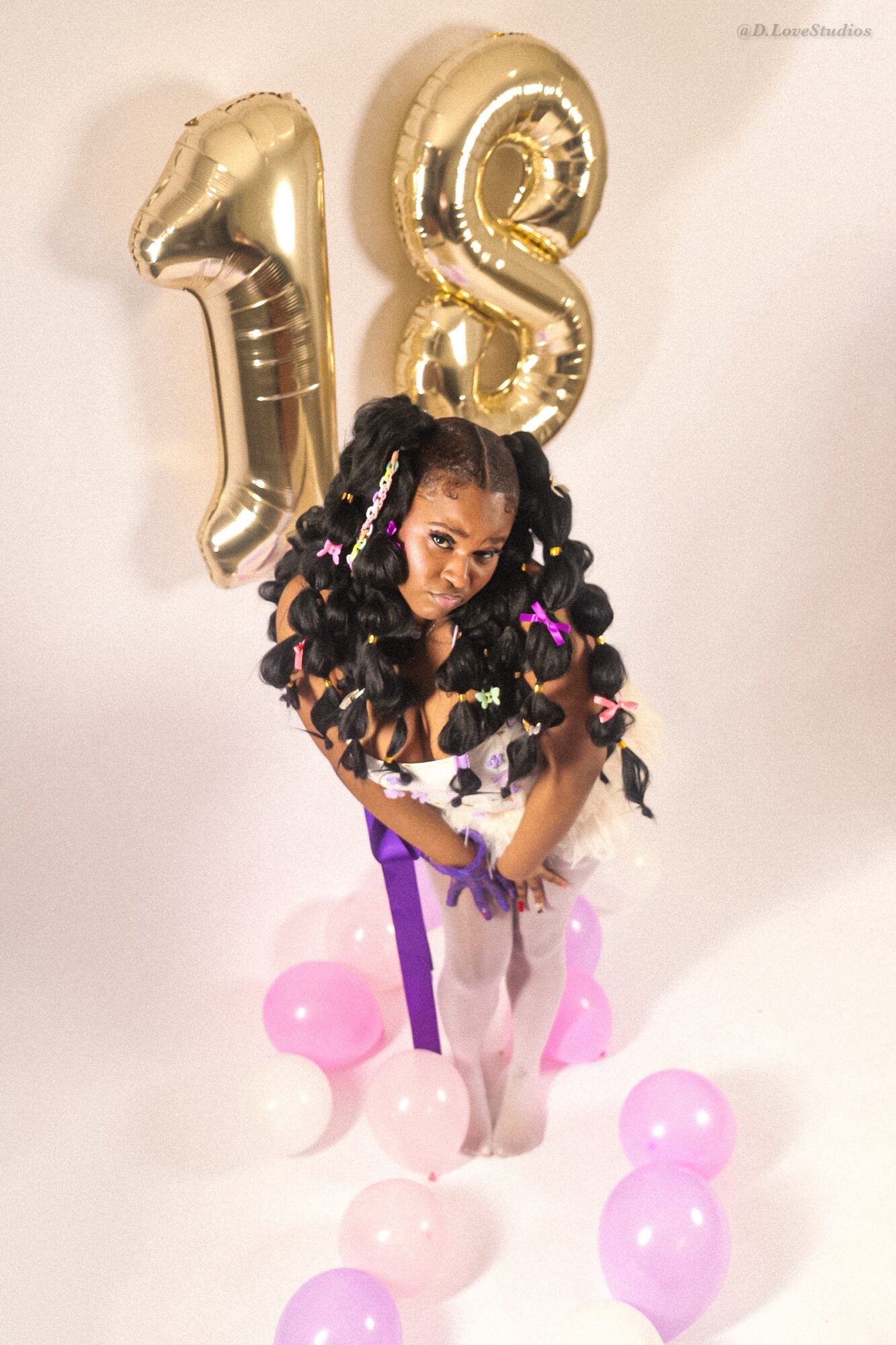 Young girl with curly hair and colorful hair accessories, standing in front of gold number balloons 1 and 8, surrounded by pink and purple balloons.