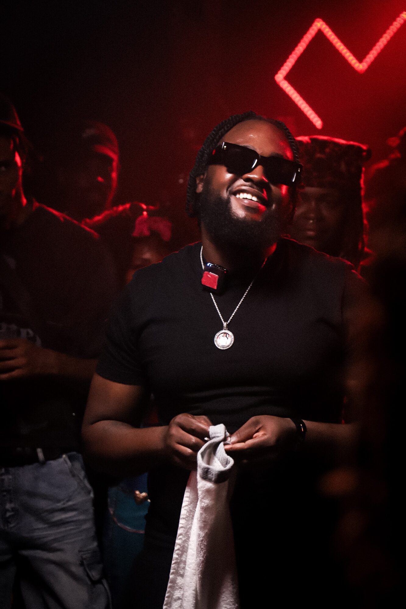 Person smiling, wearing sunglasses and a black shirt, holding a cloth, in a dark club with red lighting and a neon sign