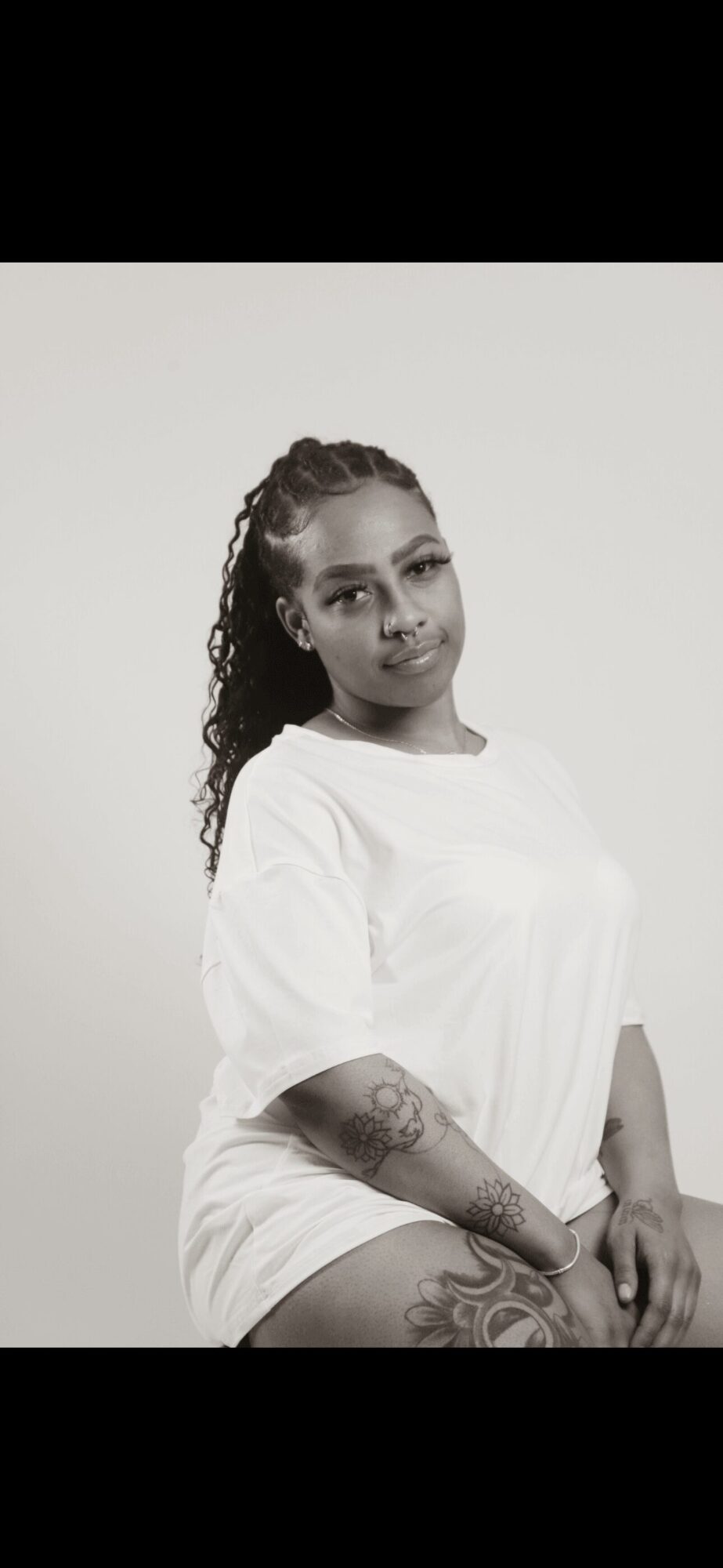Woman with curly hair and tattoos, wearing a white shirt, sitting against a plain background.