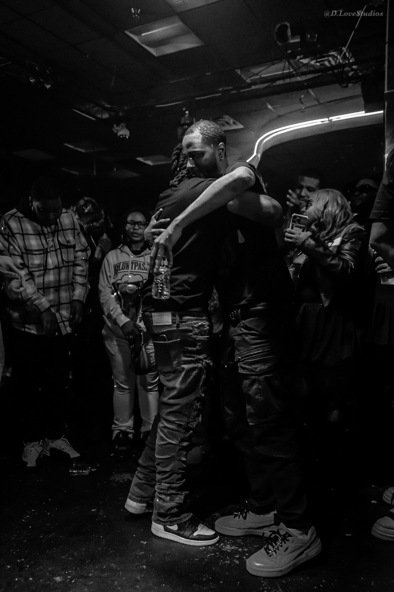 Two people hugging in a dark, crowded indoor setting with other people around, some smiling and watching.