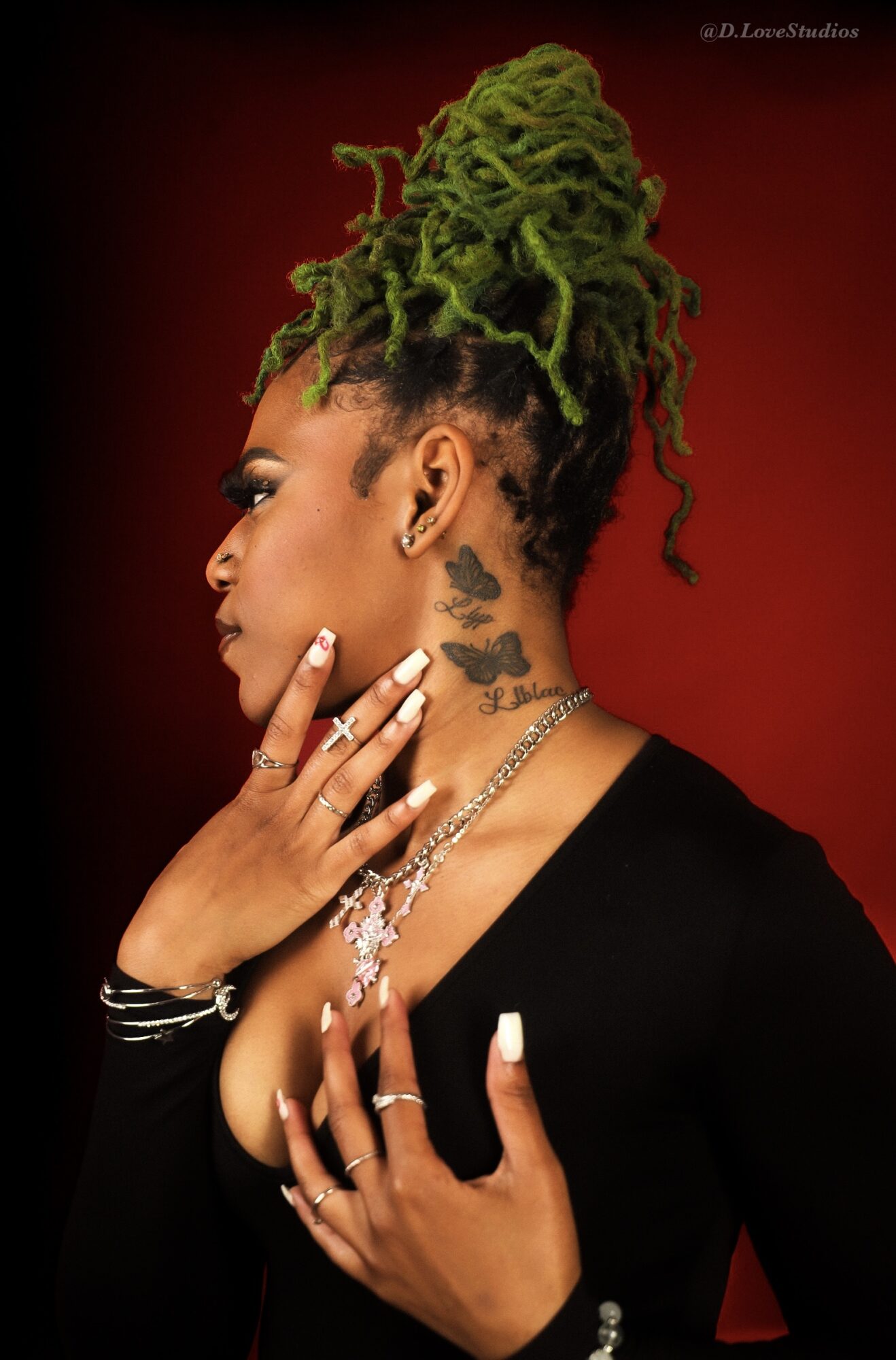 Woman with green dreadlocks styled in an updo, wearing jewelry and a black top, against a red background.