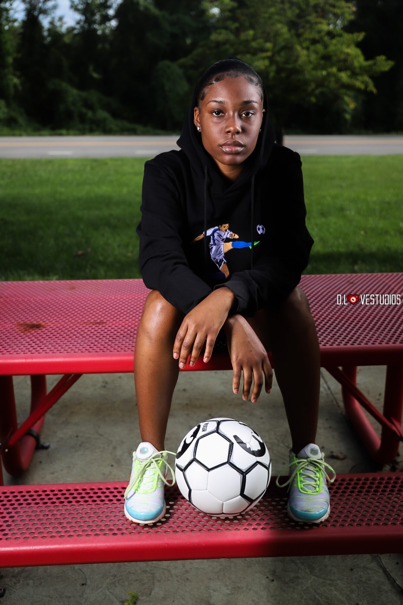 Young girl sitting on a red bench outdoors with a soccer ball, wearing a black hoodie and colorful sneakers.