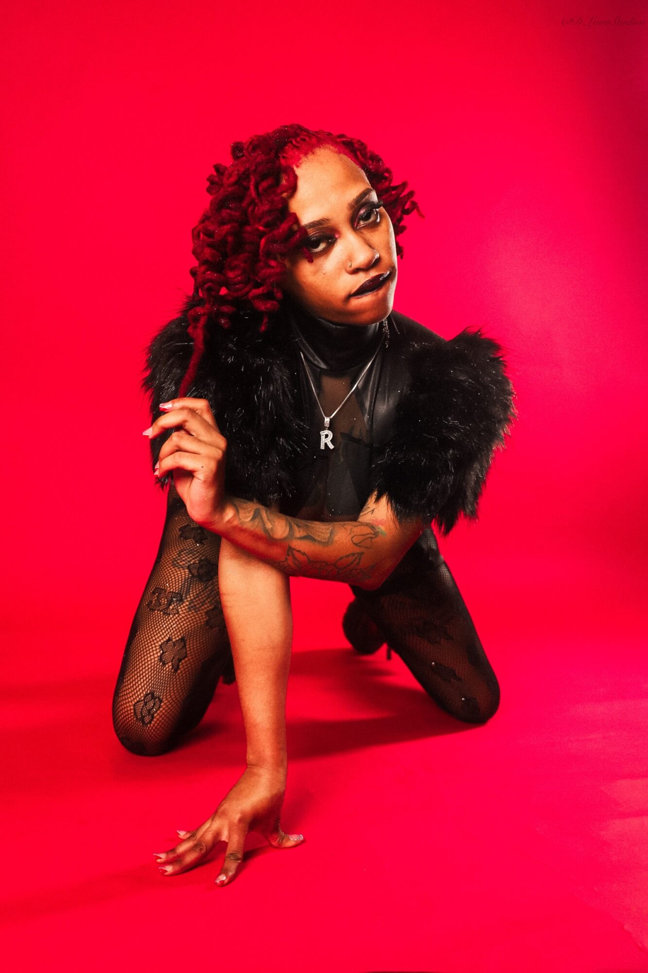 Woman with curly hair and tattoos poses on a red background, wearing black clothing and looking at the camera.