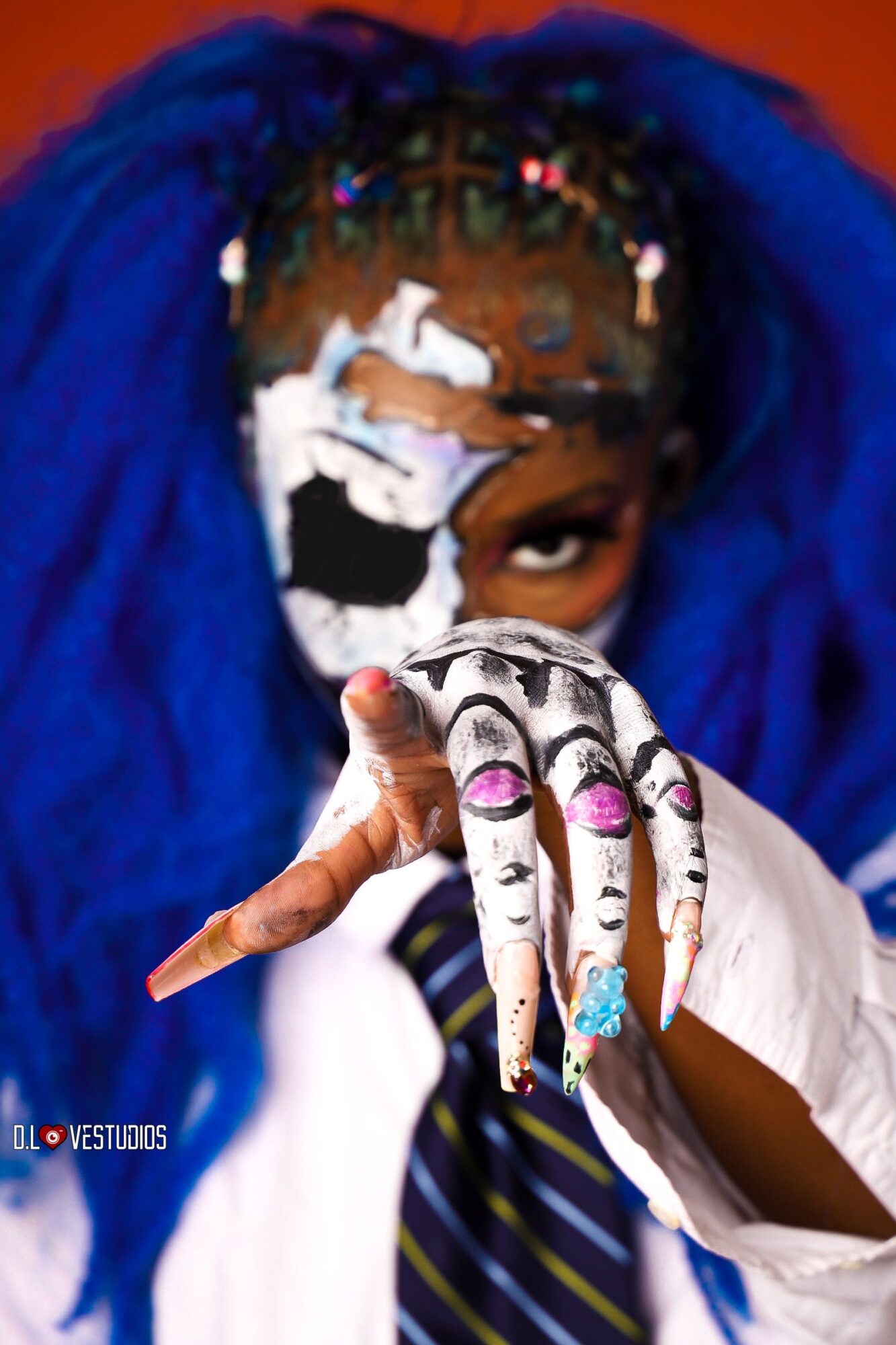 Person with blue hair and face paint, holding hand with painted fingers and rings, partially covering face, against colorful background.