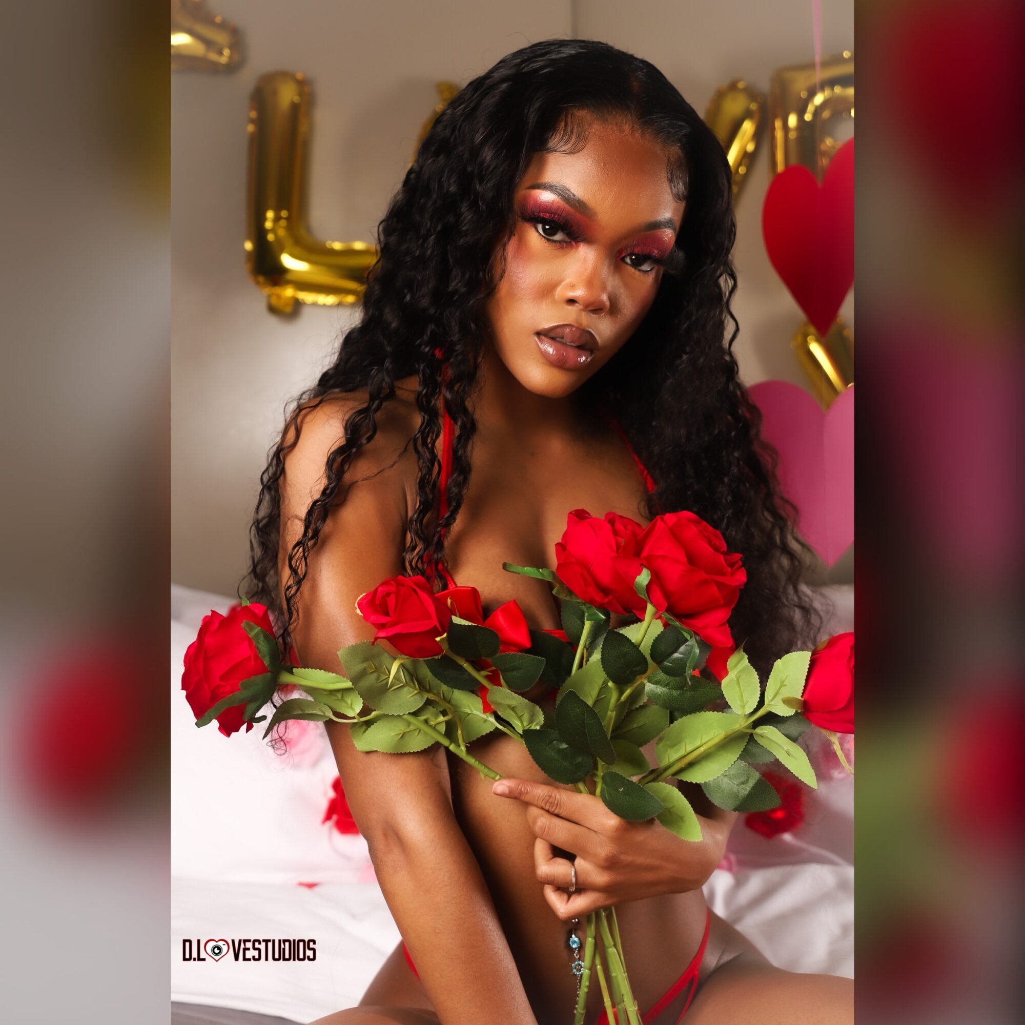 Woman with long curly hair holding a bouquet of red roses, decorated with gold and pink balloons in the background.
