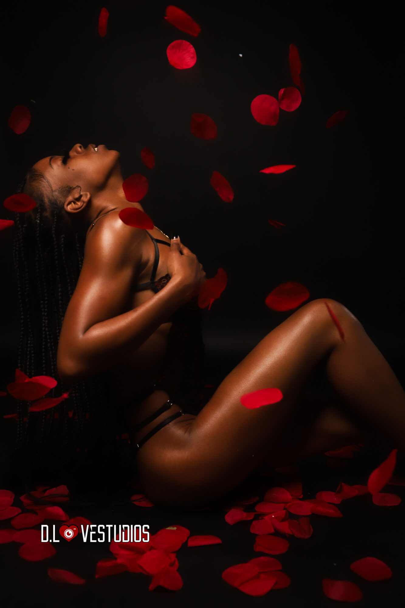 Nude woman sitting with head tilted back among red rose petals against black background.