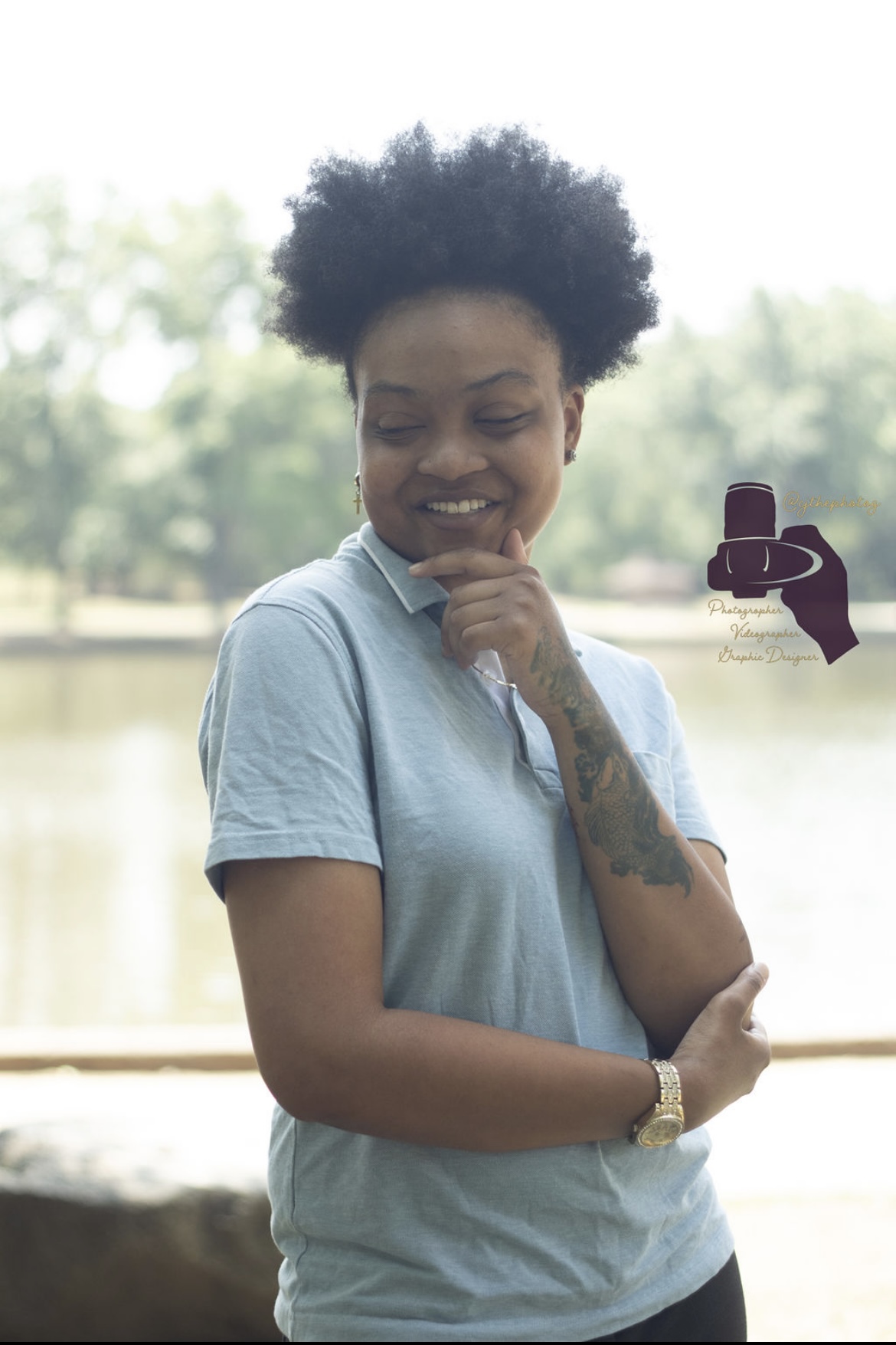 Young woman with curly hair smiling outdoors, wearing a light blue shirt, with trees and water in background.