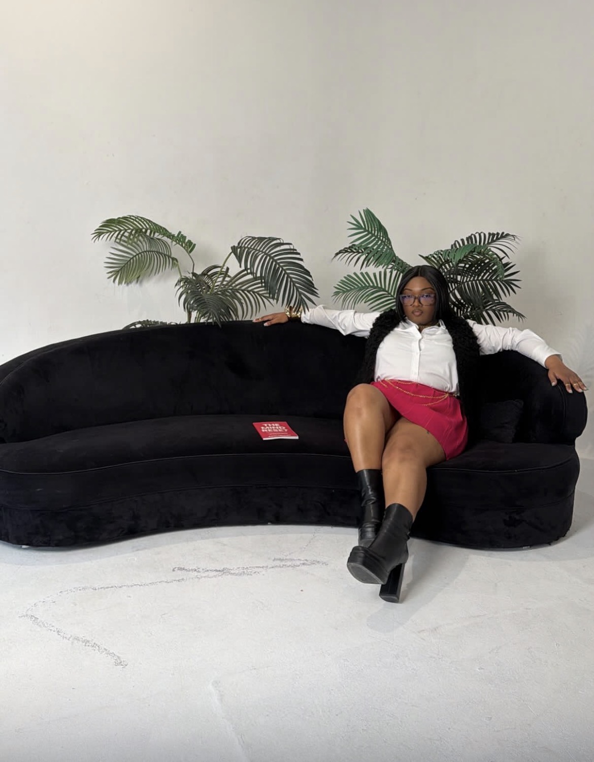 Person sitting on a black sofa with plants behind, wearing a white top, red skirt, and black boots, in a minimal room.