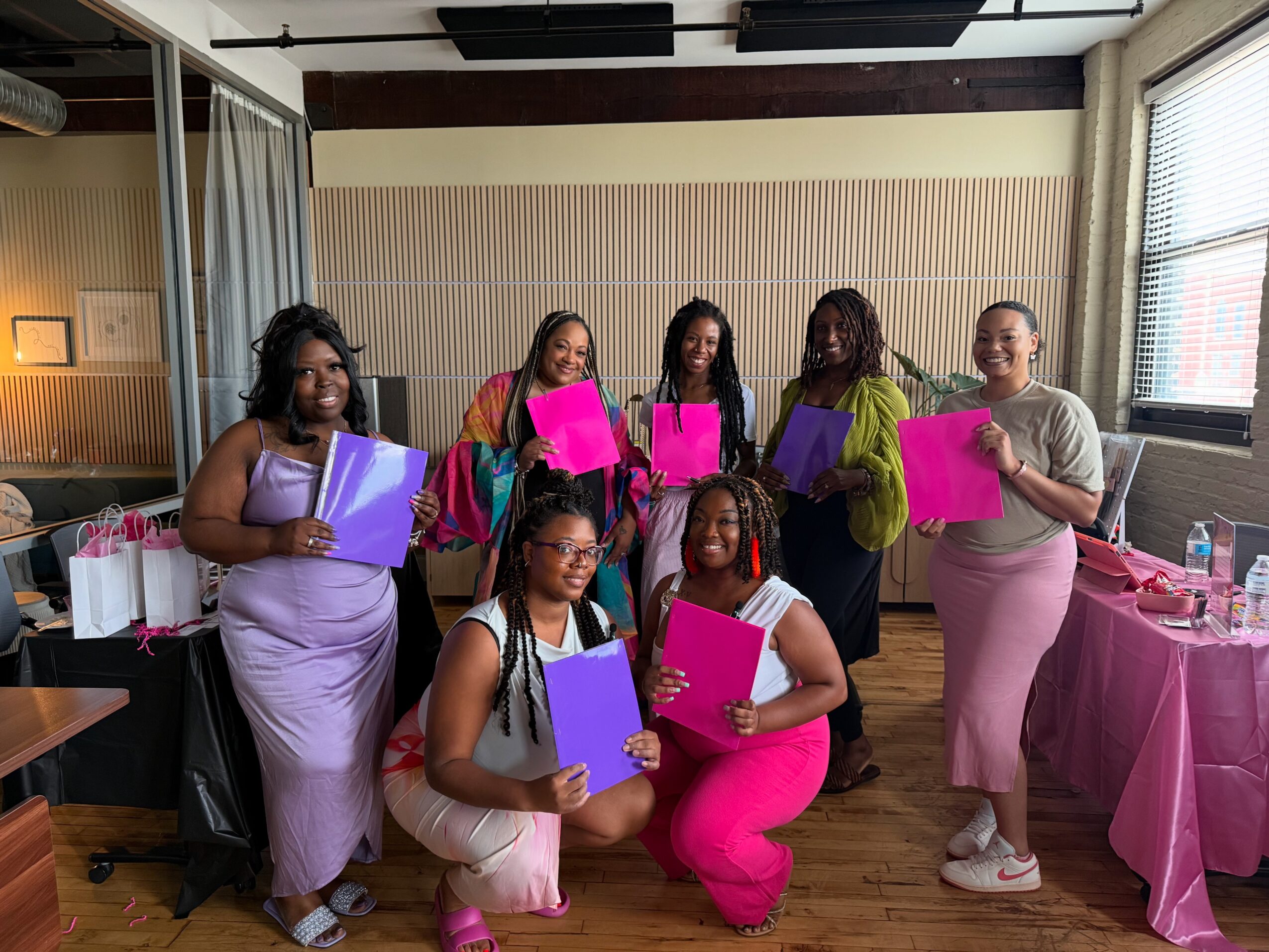 Seven women holding colorful folders and bags, standing and kneeling indoors with a pink table and window in background.