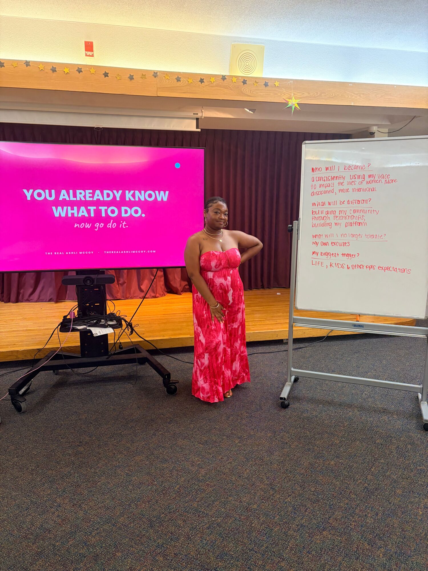 Woman in red dress standing on stage with a microphone, whiteboard and pink screen behind her.