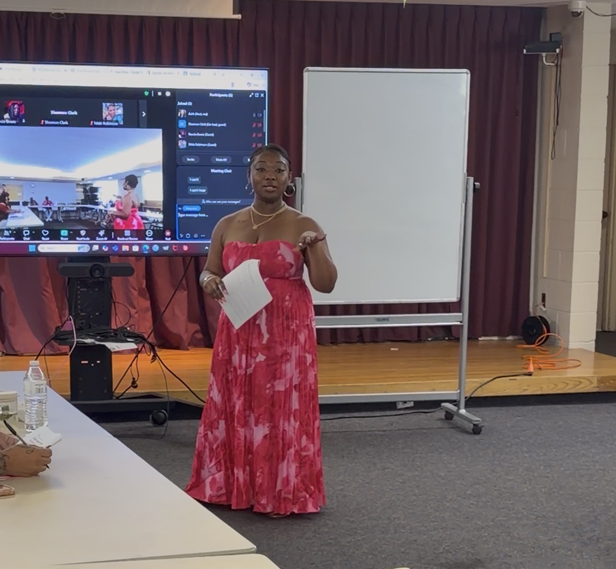Woman in a pink dress holding papers, standing in front of a whiteboard and screen in a conference room.