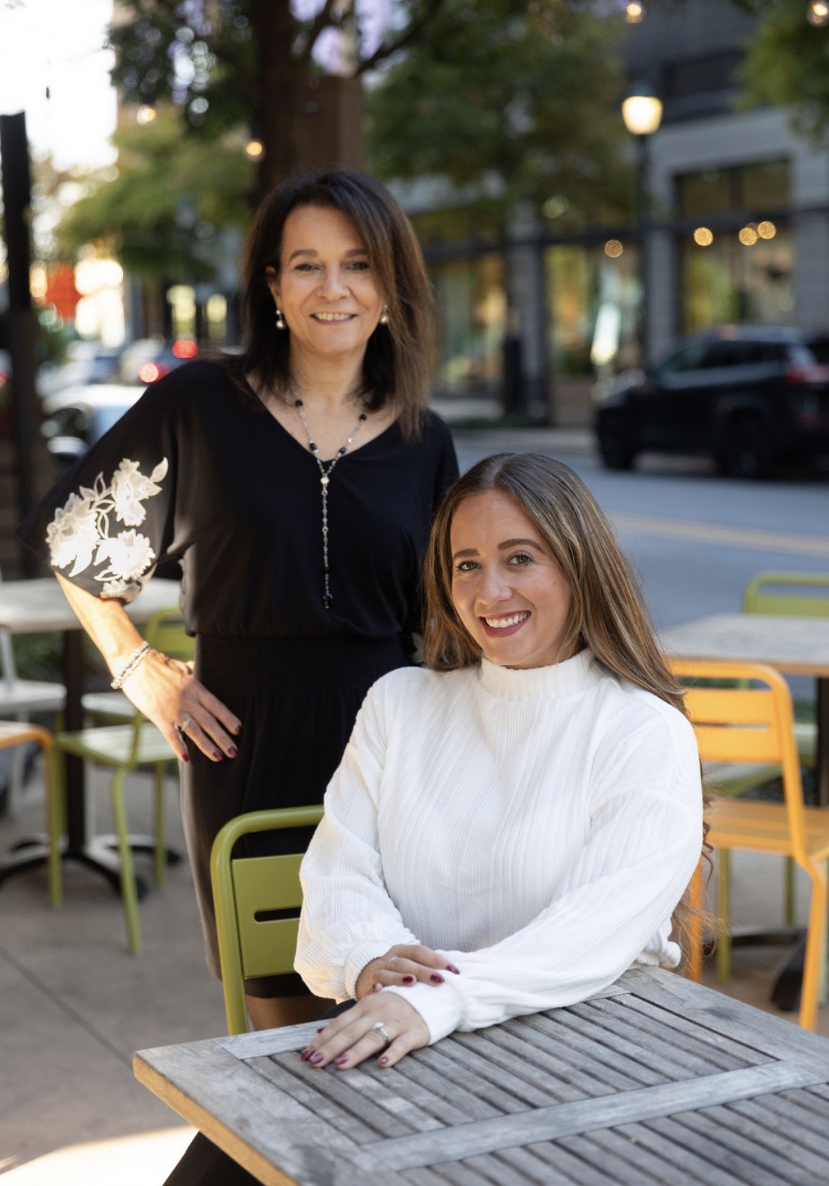 Two women smiling outdoors at a table with colorful chairs, trees, and buildings in the background.