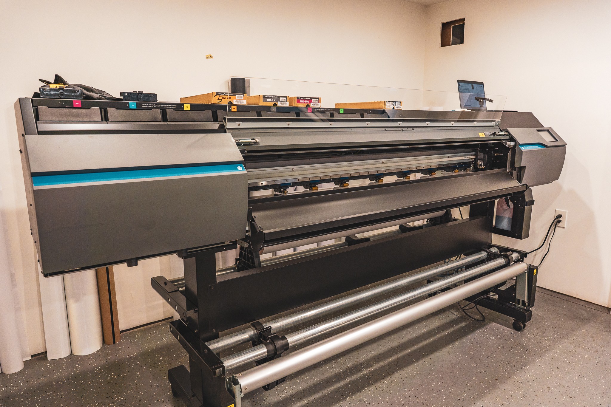 Large industrial printer with multiple rollers and control panel, positioned against a white wall in a room.