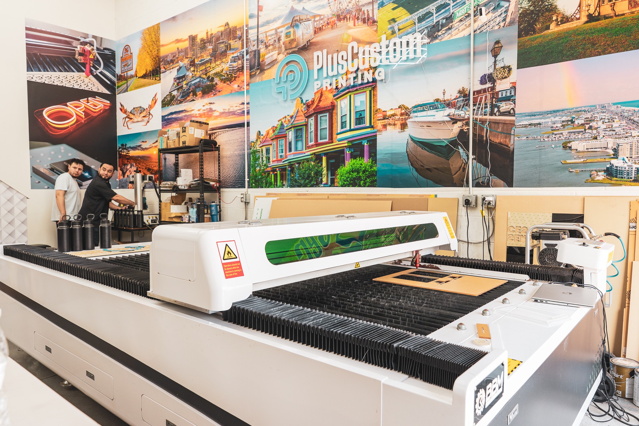 Large laser cutting machine in workshop with colorful cityscape mural on wall behind, and a person working nearby.