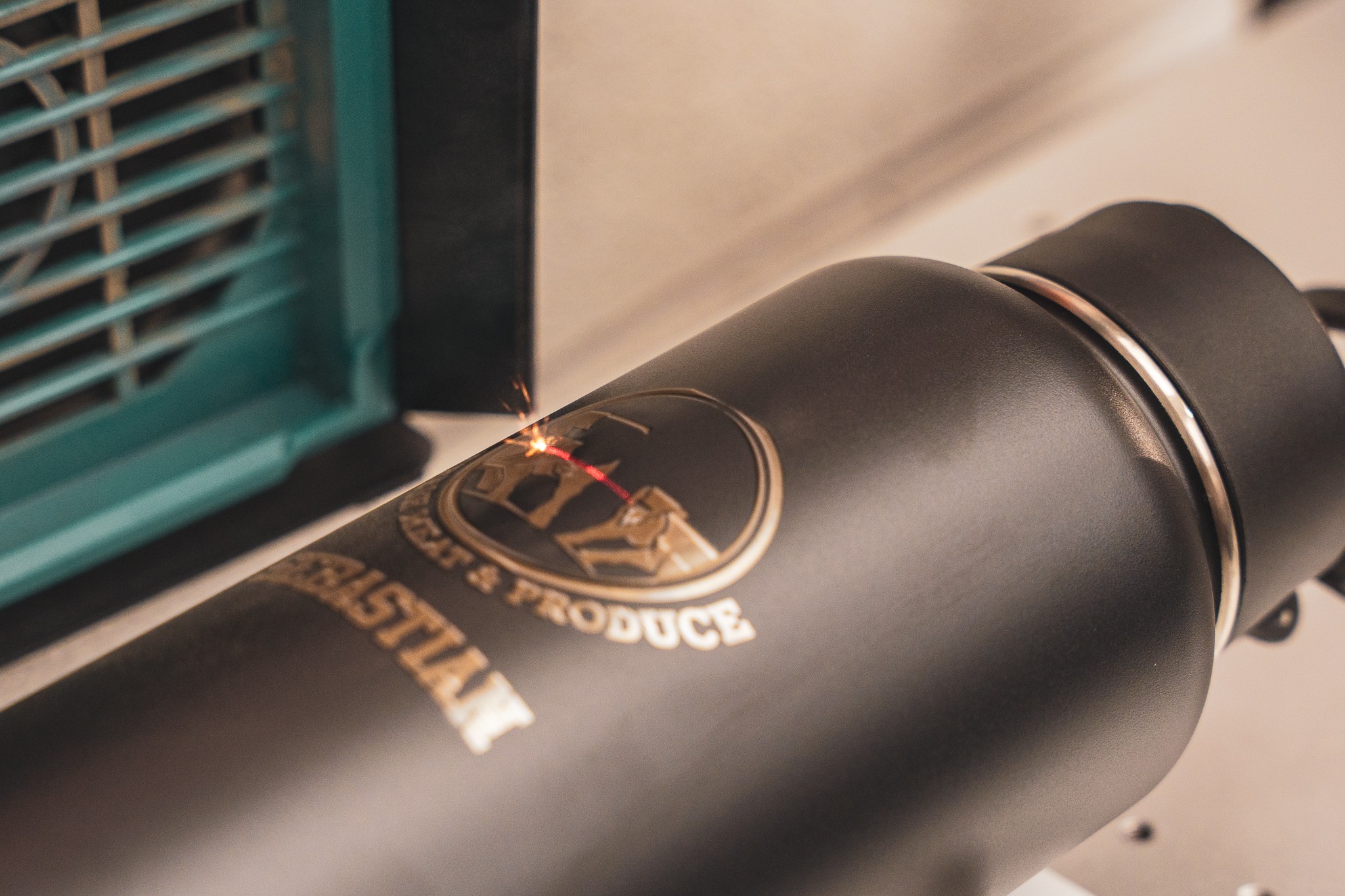 A black cylindrical object with a logo being welded or engraved, with a green vent or grill nearby, on a beige surface.