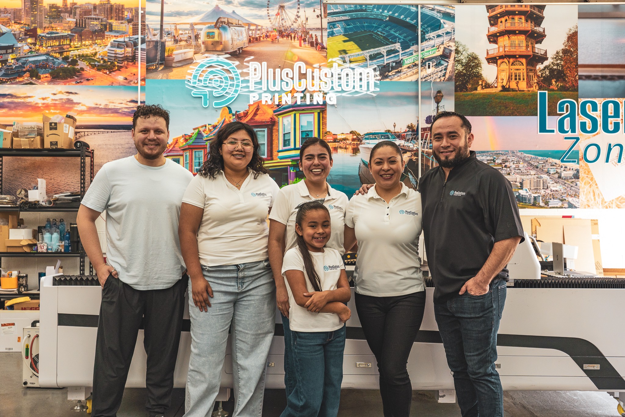 Group of six people standing in front of a colorful sign with cityscape images, smiling, indoors.