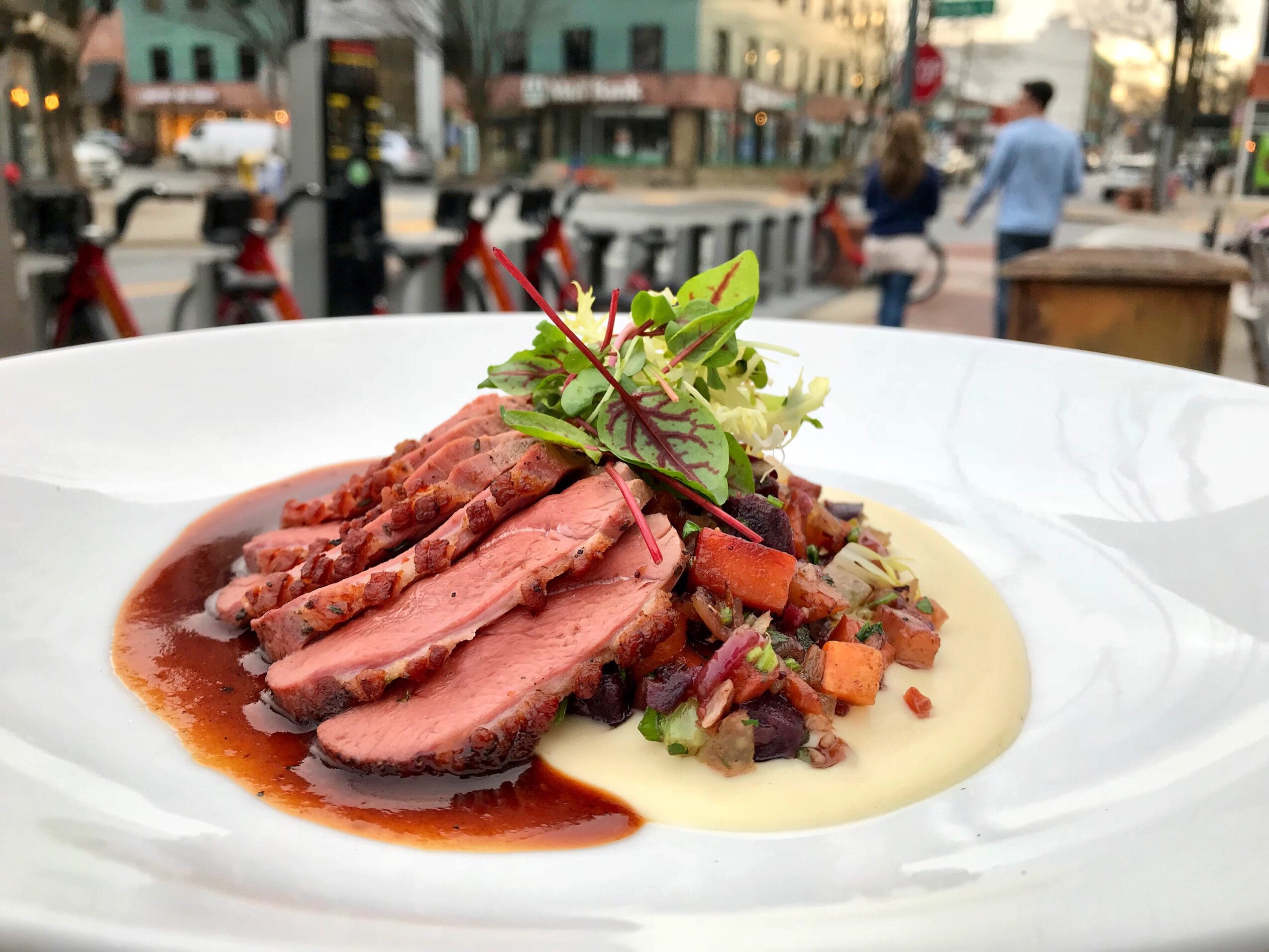 Slices of cooked meat with sauce, garnished with greens, served with a side of vegetables and mashed potatoes on a white plate
