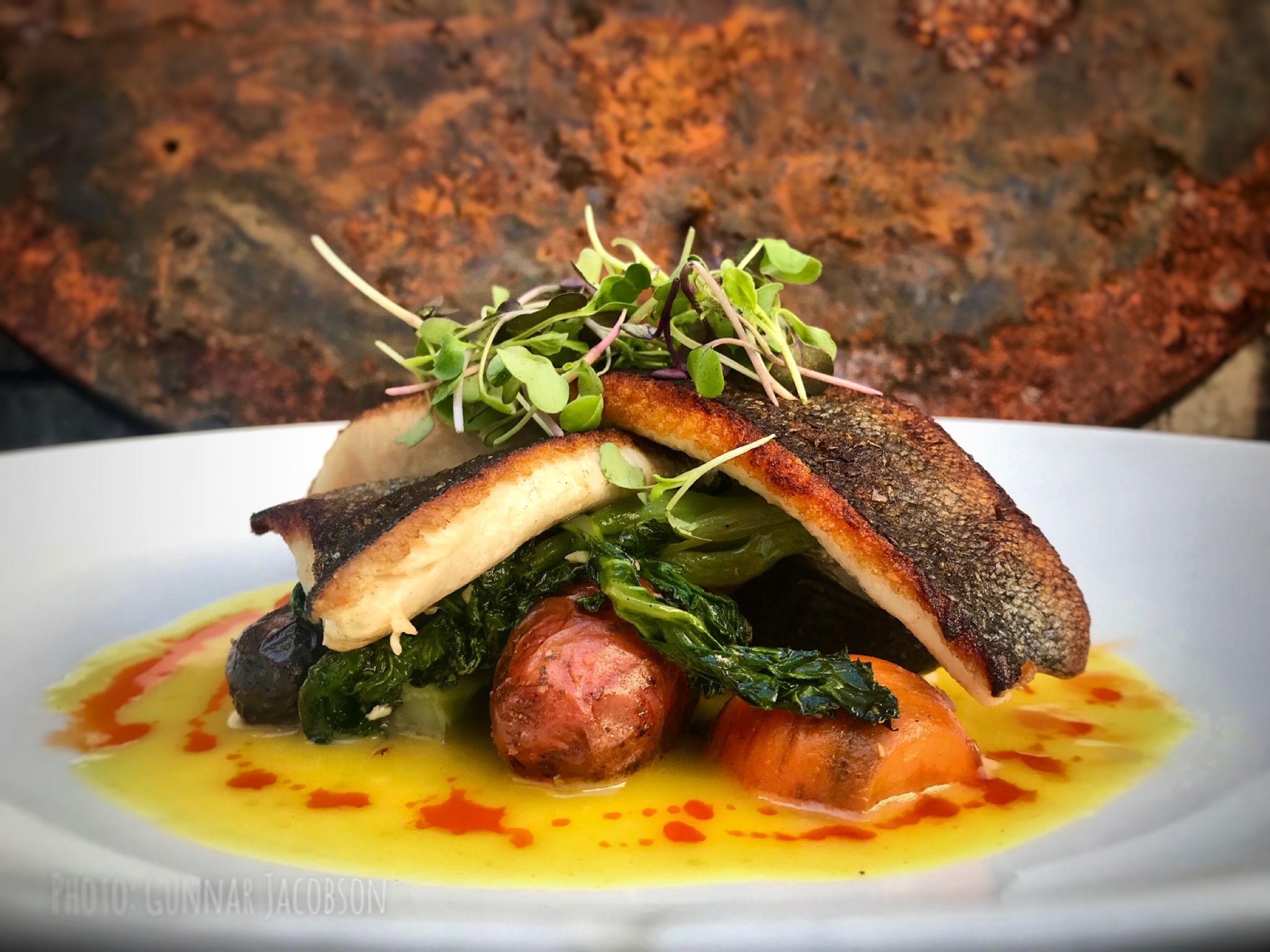 Grilled fish fillet with greens, sausage, and vegetables on a white plate, garnished with microgreens, with a rust-colored background.