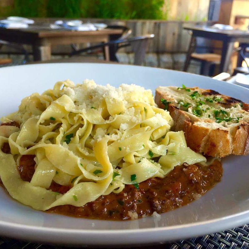 Plate with pasta, meat sauce, and garlic bread, set outdoors with wooden tables and chairs in background.