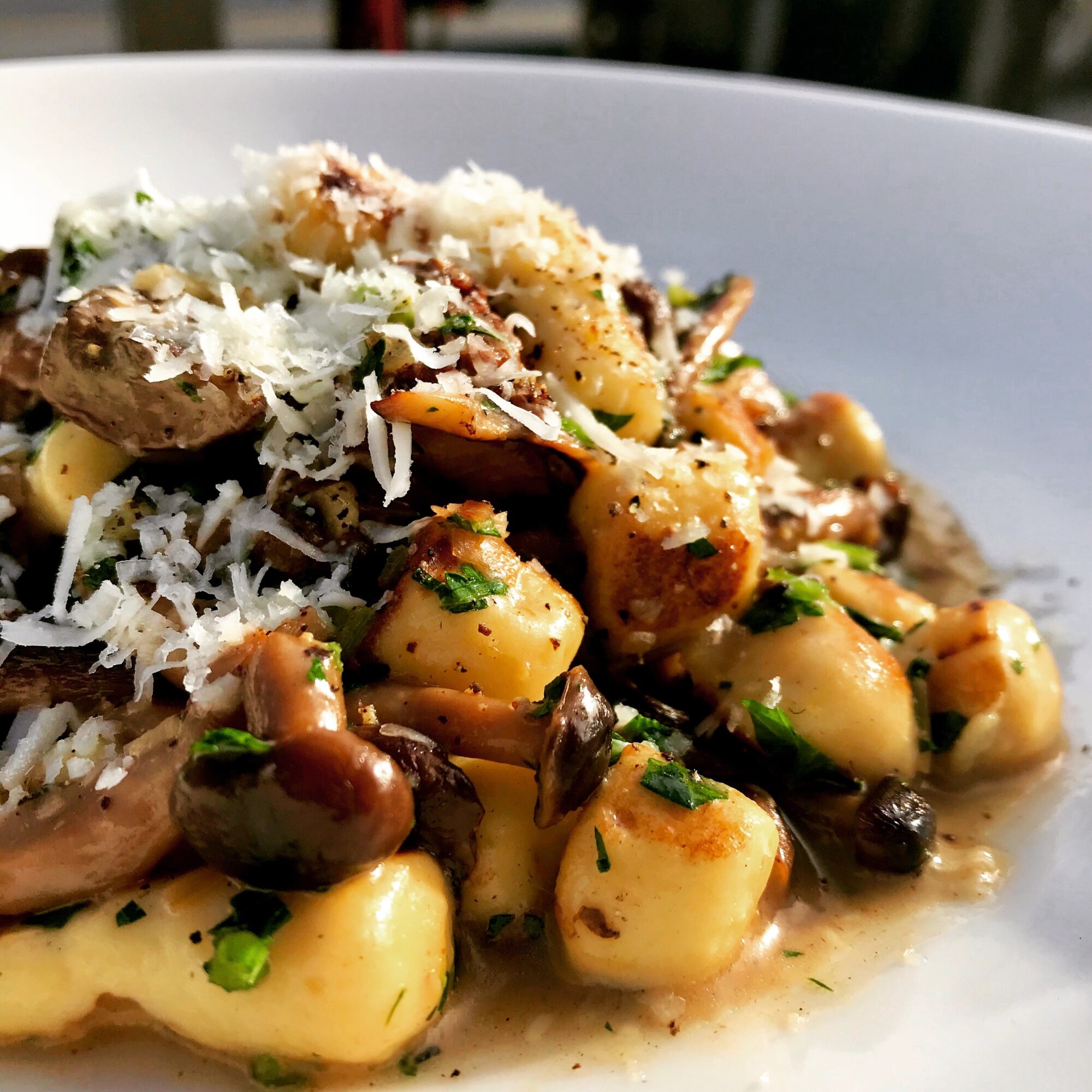 Close-up of gnocchi with mushrooms, herbs, and grated cheese on a white plate, outdoors, natural light.