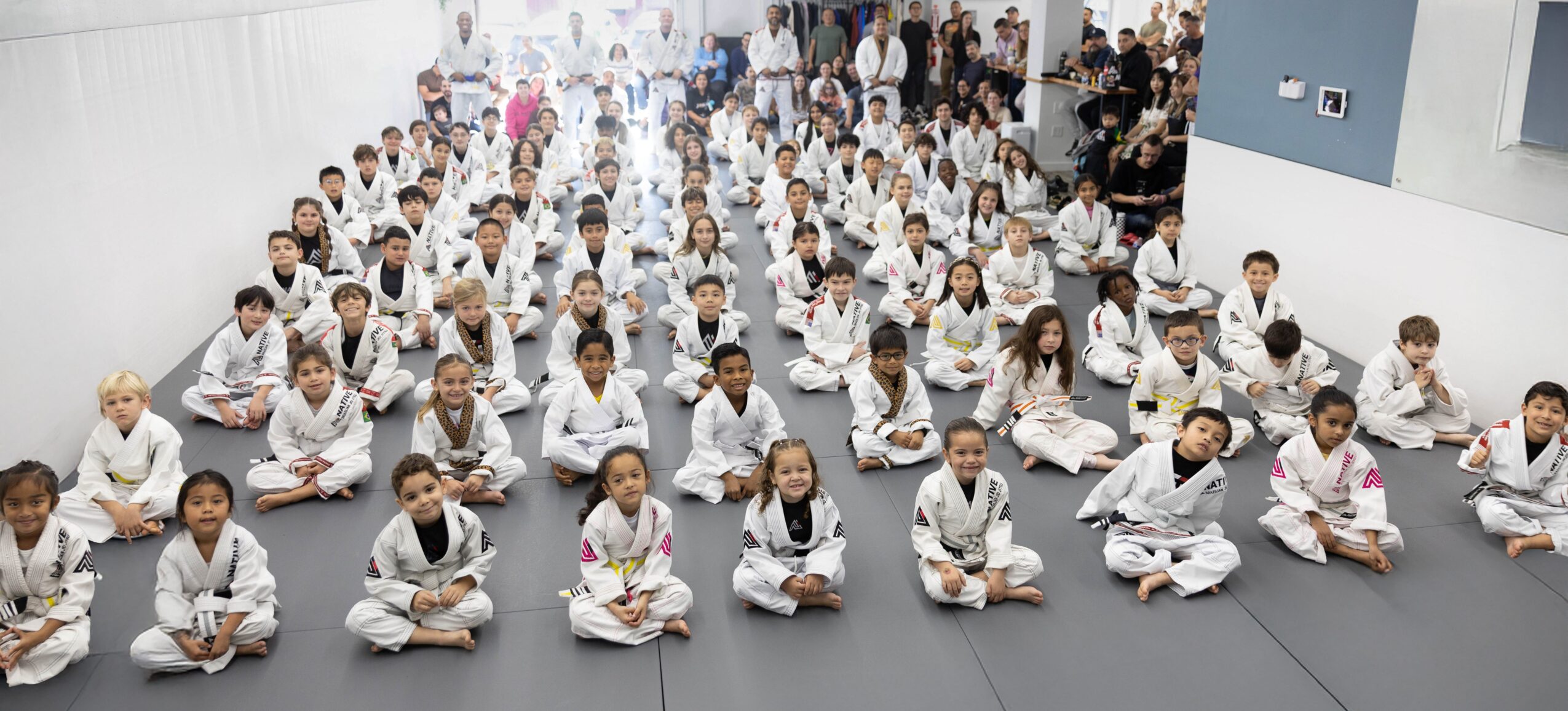 Large group of children sitting cross-legged on mats in a spacious room, facing forward, wearing white uniforms.