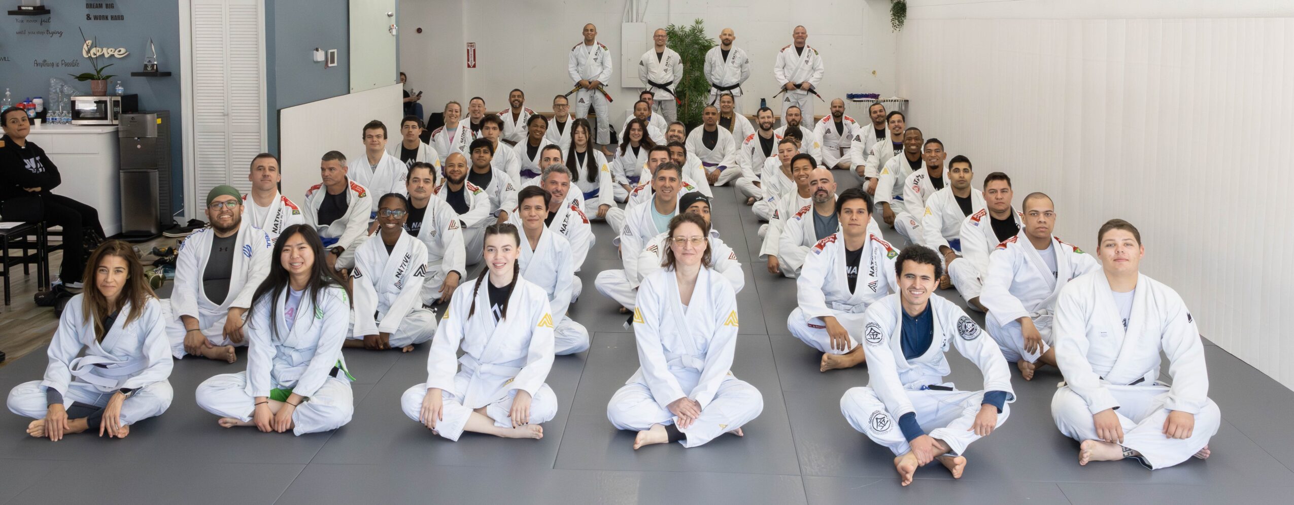 Group of people in white uniforms sitting on the floor in a spacious room, facing forward, with some standing in the background.