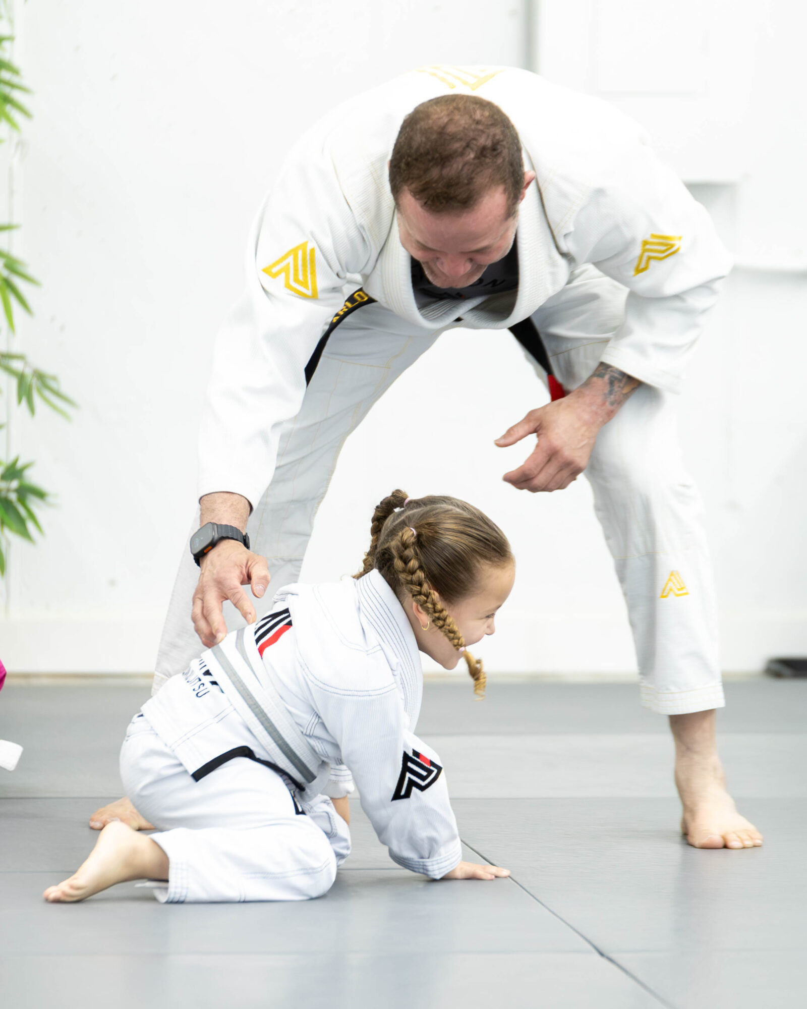 A person in a white martial arts uniform helps a young girl in a similar uniform on the floor during training.