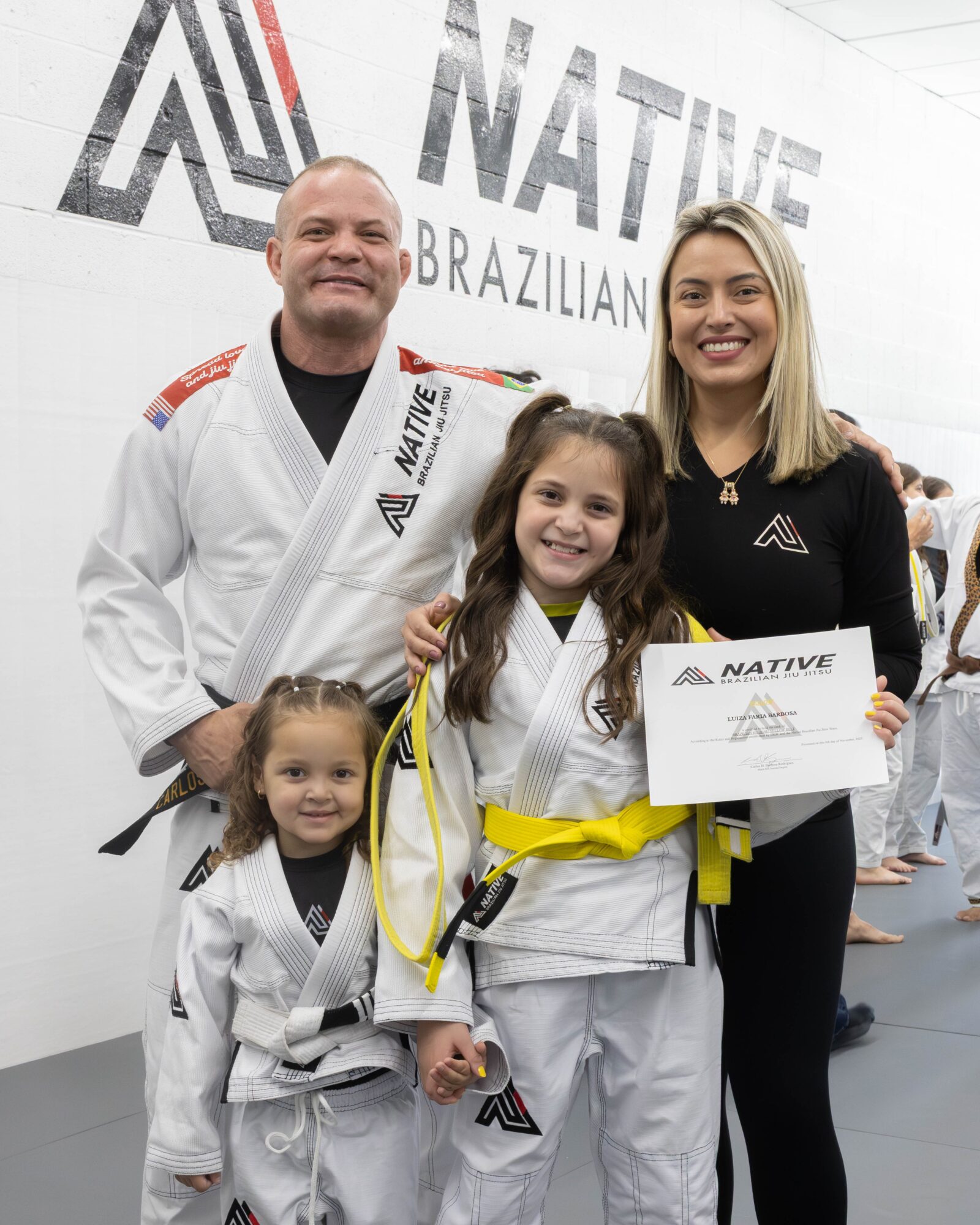 Two adults and two children in Brazilian Jiu-Jitsu uniforms smiling indoors, holding a certificate, with a logo and text in the background.