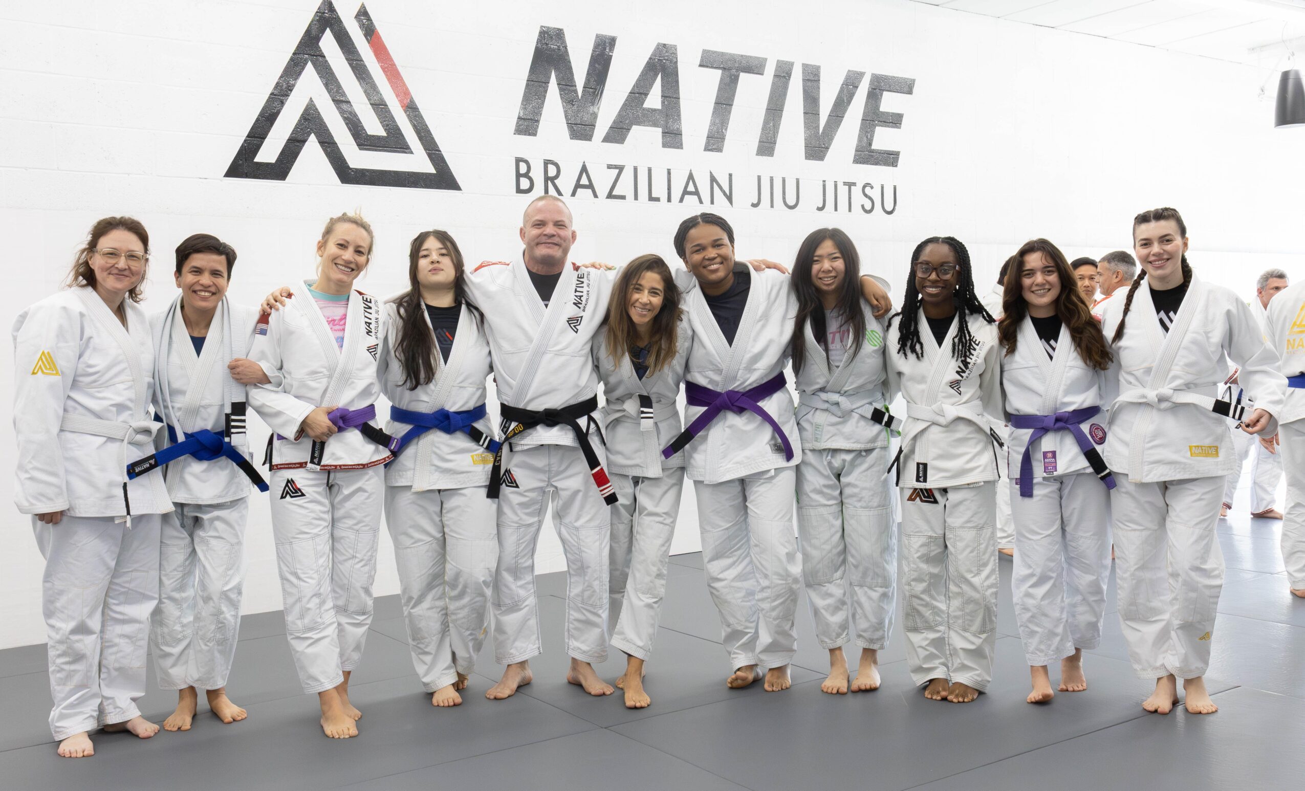 Group of people in Brazilian Jiu Jitsu uniforms standing together, smiling, in front of a banner with the words 'NATIVE BRAZILIAN JIU JITSU'.