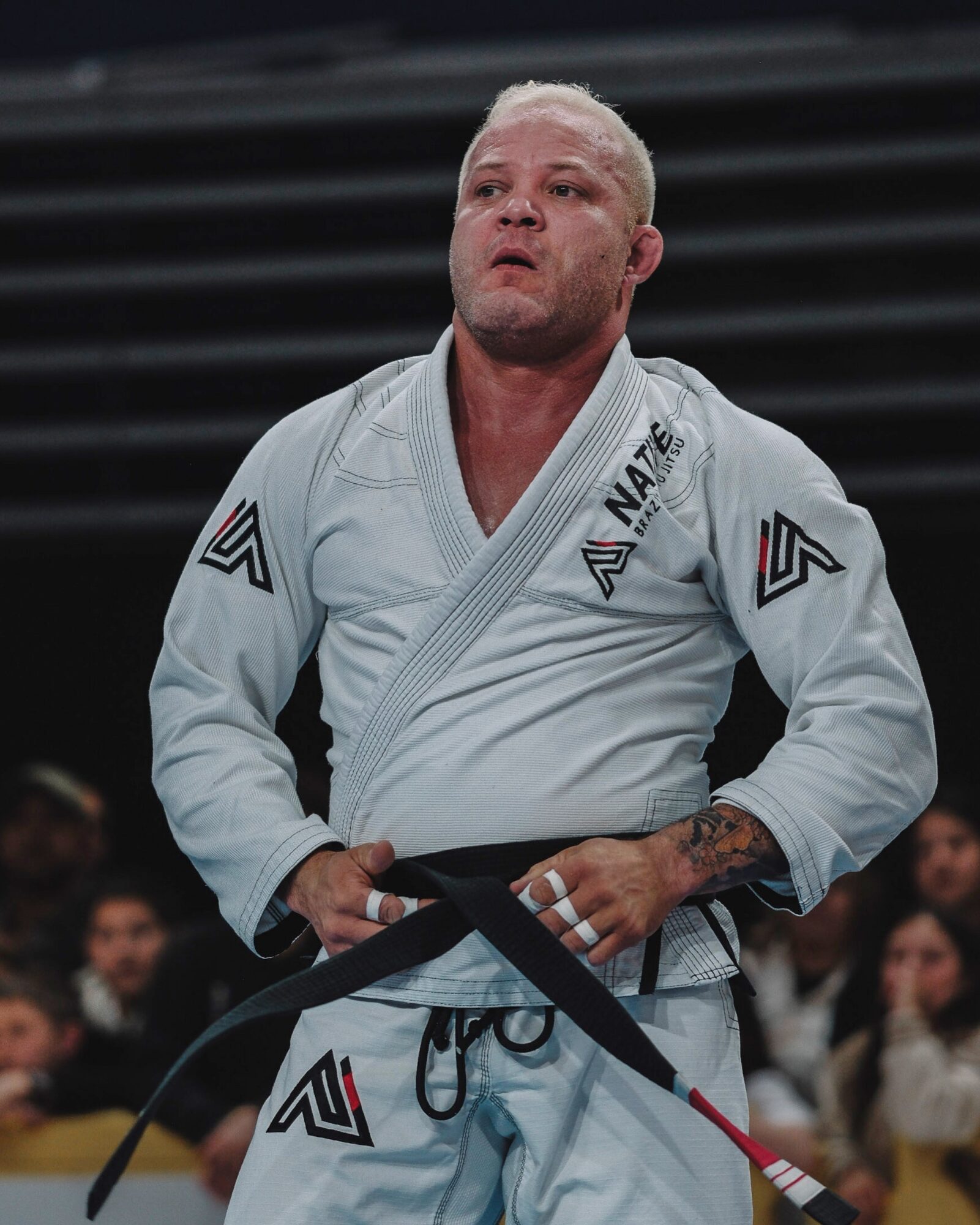 Person in a white martial arts gi with black belt, standing in a gym or competition setting, holding a red and black belt.