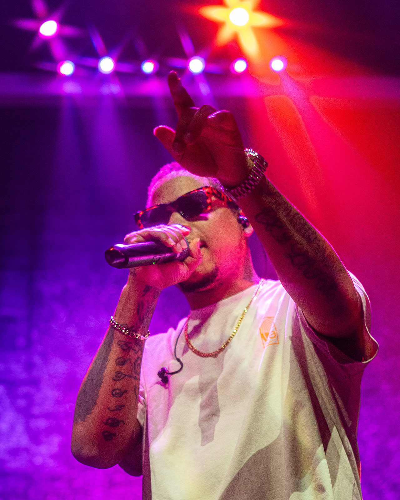 Performer with sunglasses singing into a microphone on stage, colorful lights overhead, wearing a white shirt and jewelry.