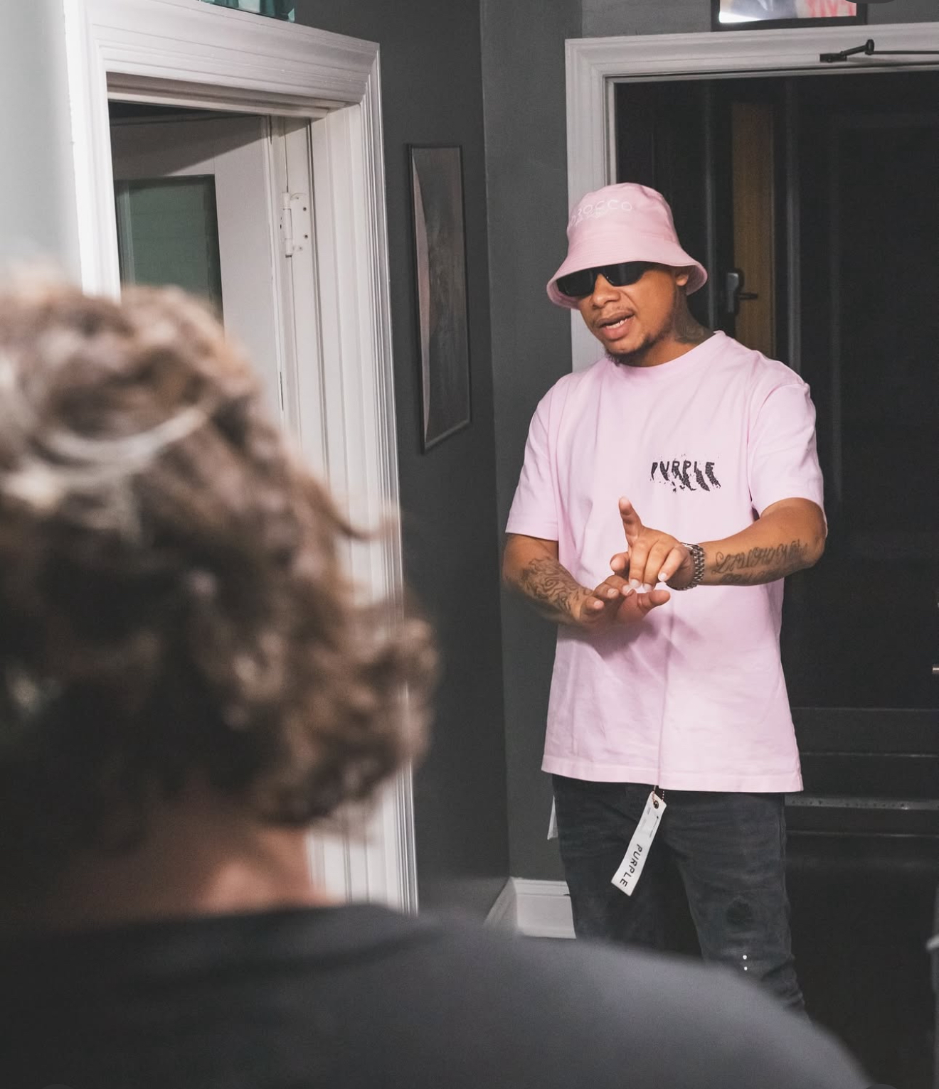 Person wearing pink bucket hat, sunglasses, and pink T-shirt standing indoors, talking to someone with curly hair, in a room with dark walls.