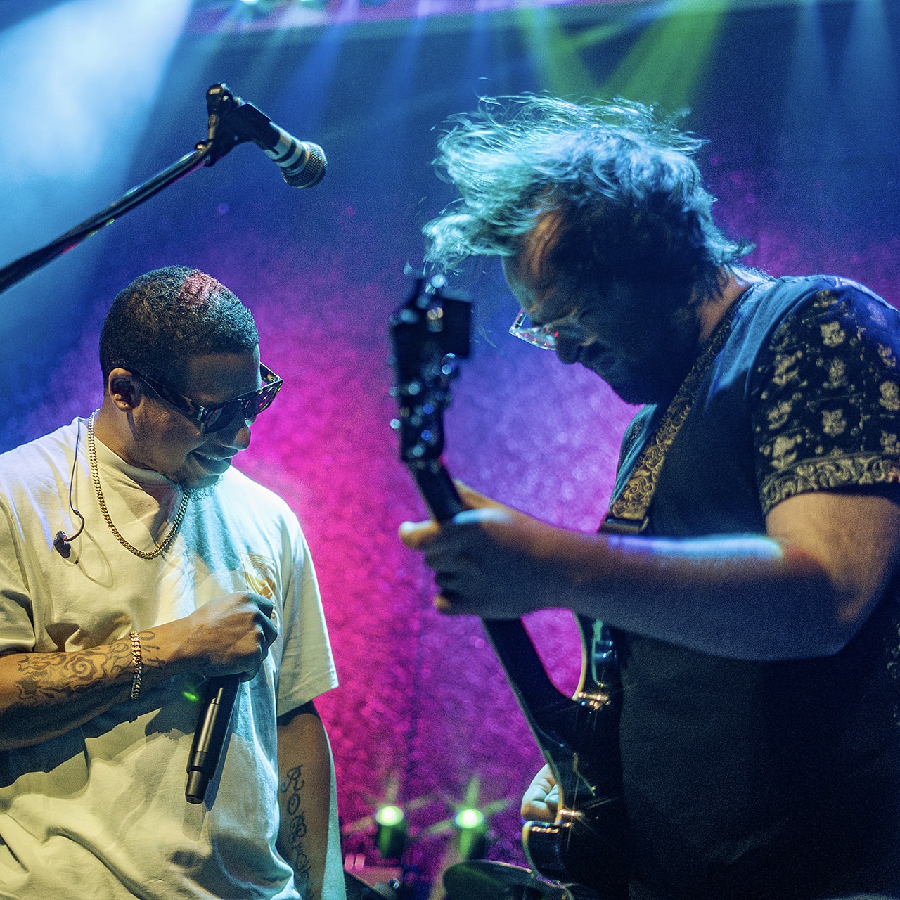 Two musicians perform on stage with colorful lighting, one holding a microphone and the other playing a guitar.
