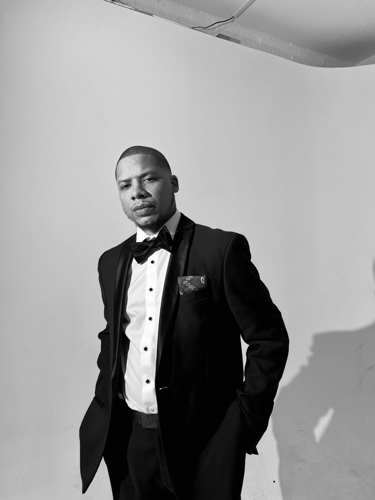 Young man in tuxedo with bow tie, standing against a plain background, looking at the camera.