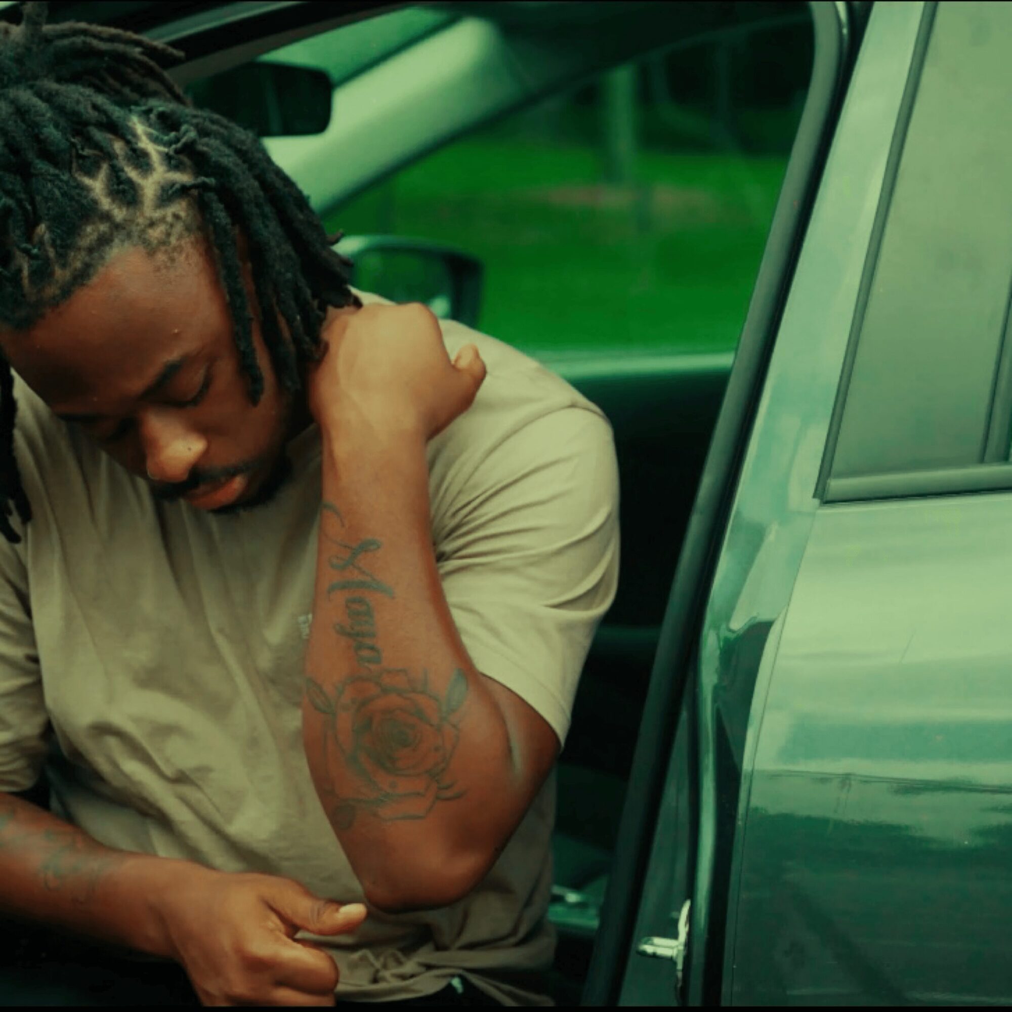 Person with dreadlocks sitting in a car, resting their head on their shoulder, showing a tattoo on their arm.