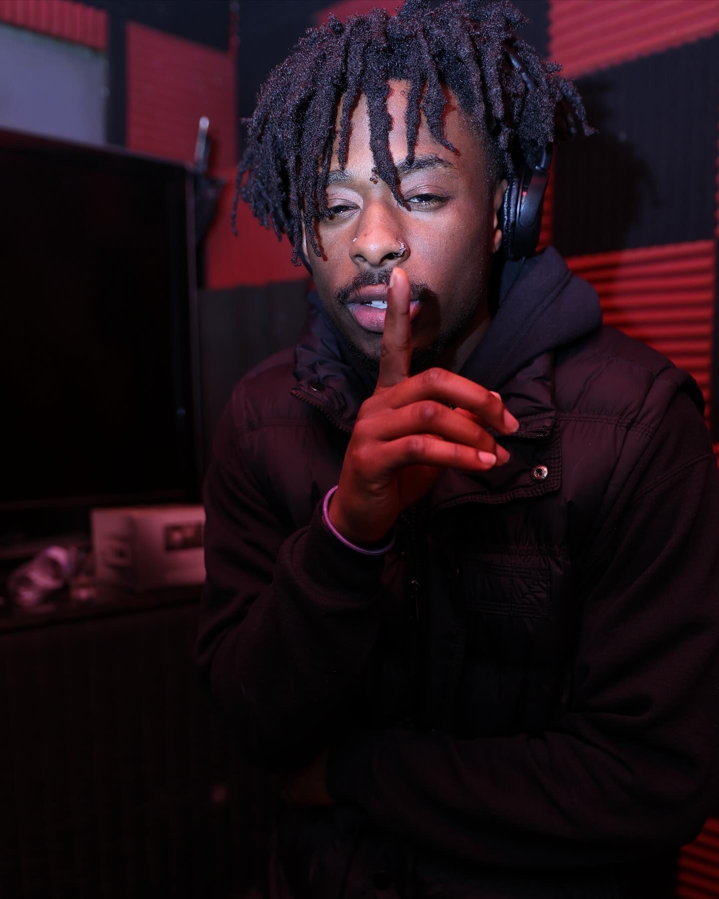 Young man with dreadlocks wearing headphones, holding finger to lips, in a recording studio with red and black walls.