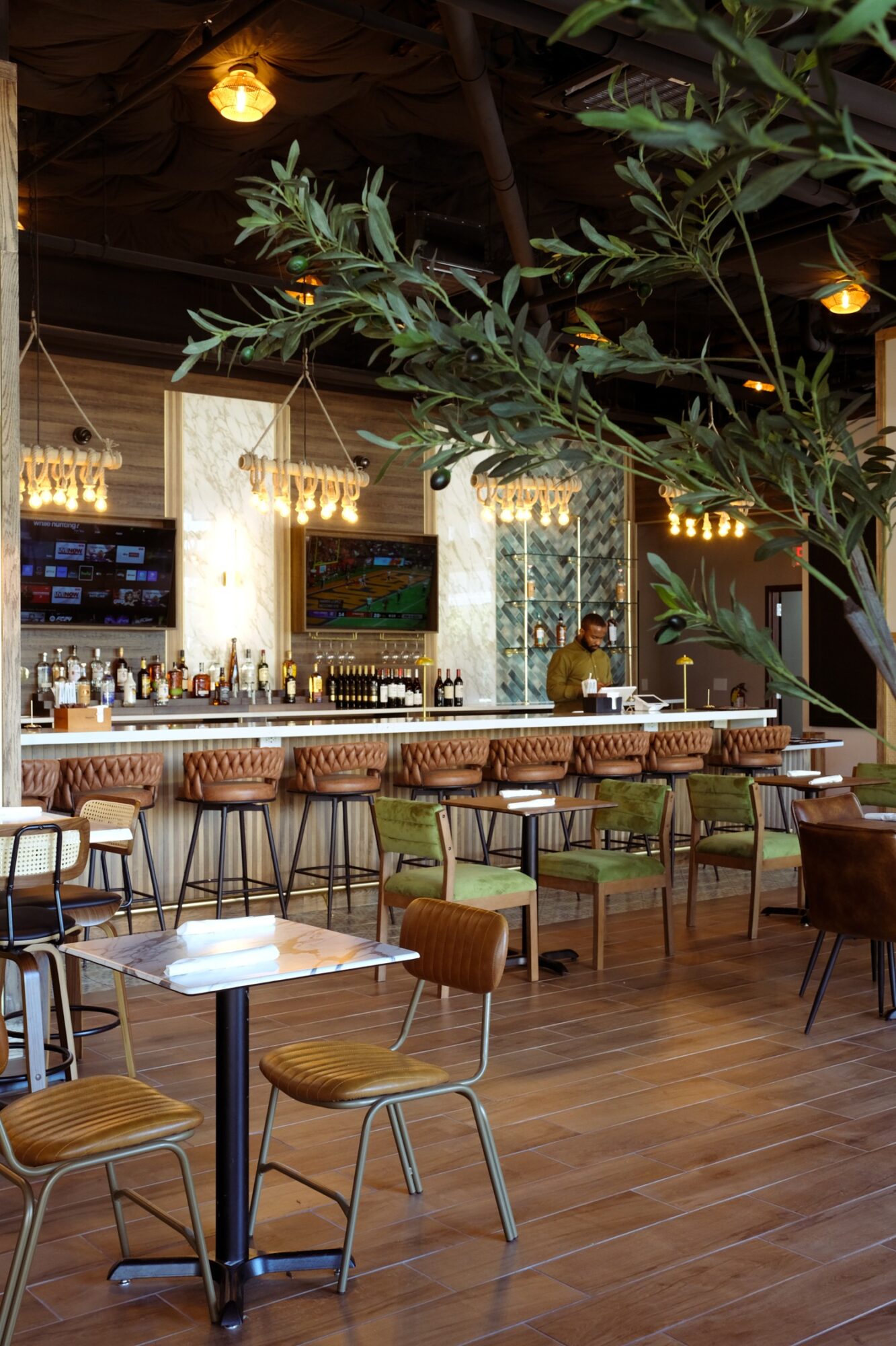 Interior of a bar or restaurant with a long counter, bar stools, tables, and chairs, decorated with hanging lights and greenery.